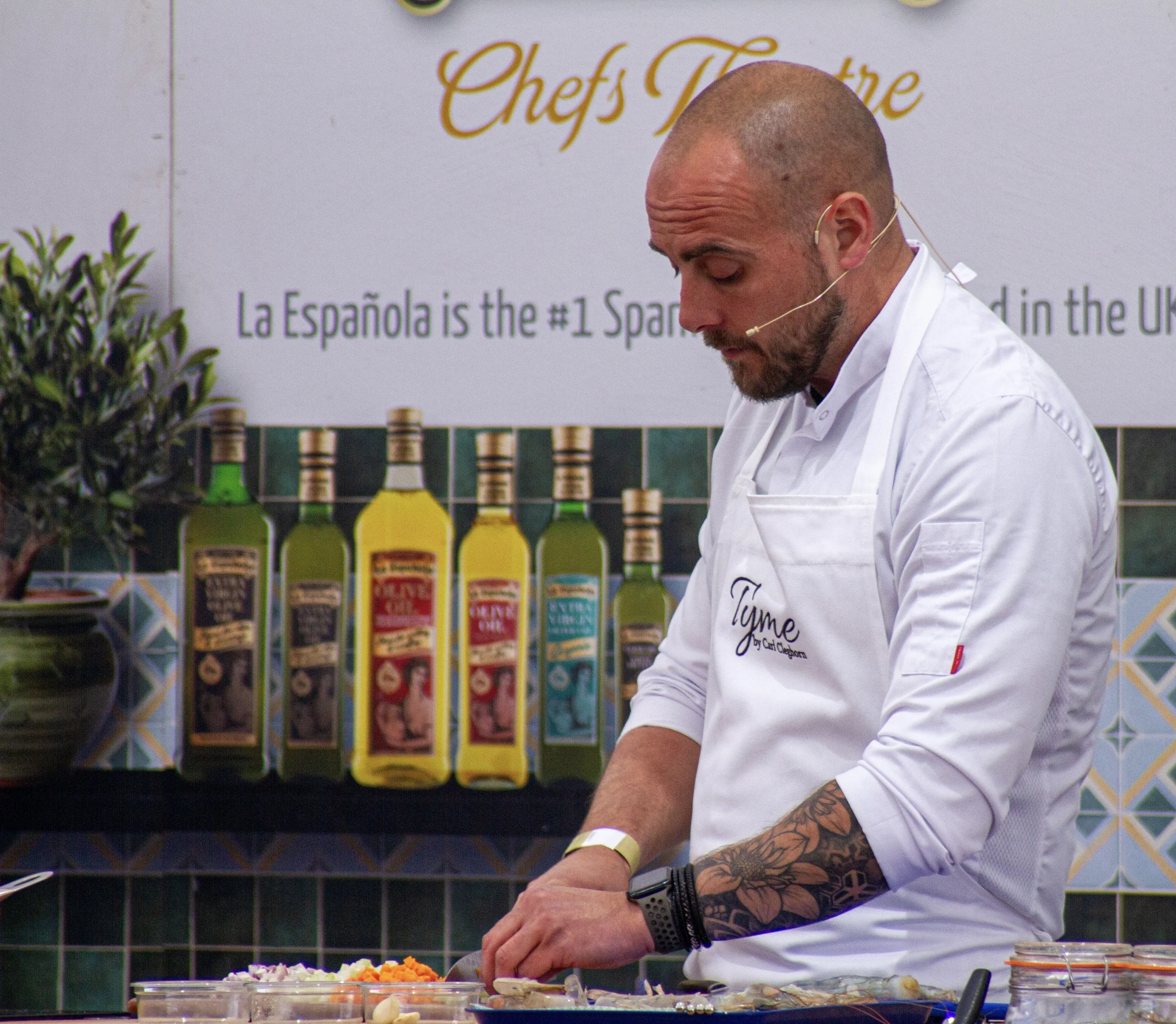 A man chef carl with a shaved head and tattoos on his left arm, wearing a white chef's coat and an apron, is preparing food at a cooking demonstration. Behind him is a sign with the words "Chef's Theatre" and bottles of cooking oils and sauces.