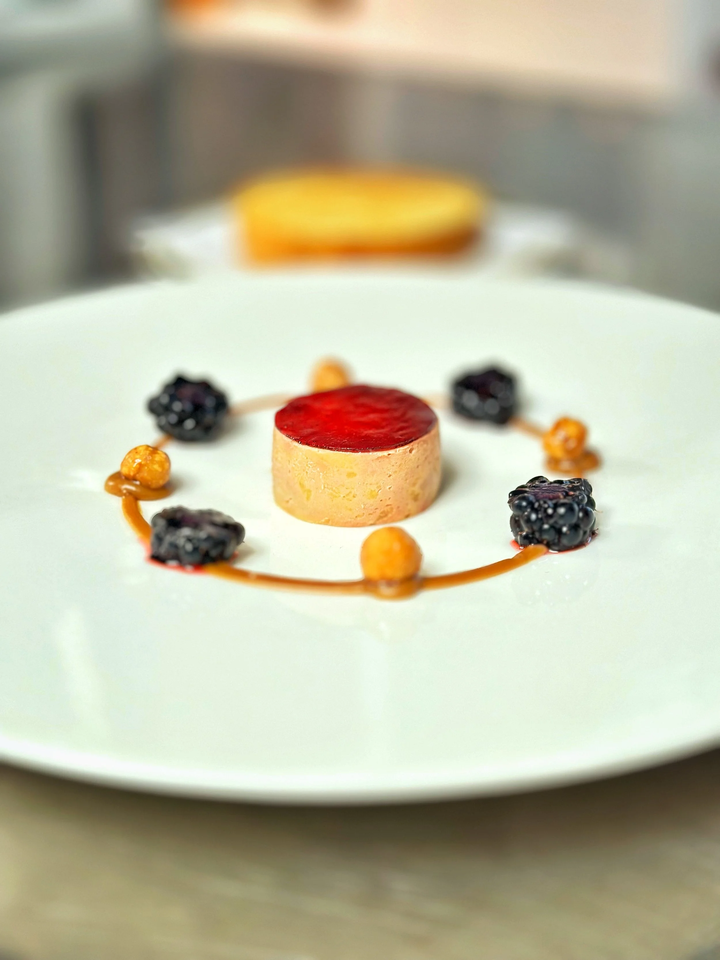 A plated duck parfait circle with blackberry sheet jelly on top, garnished with a gingerbread puree around the outside, with hazelnut and blackberry on a white plate.