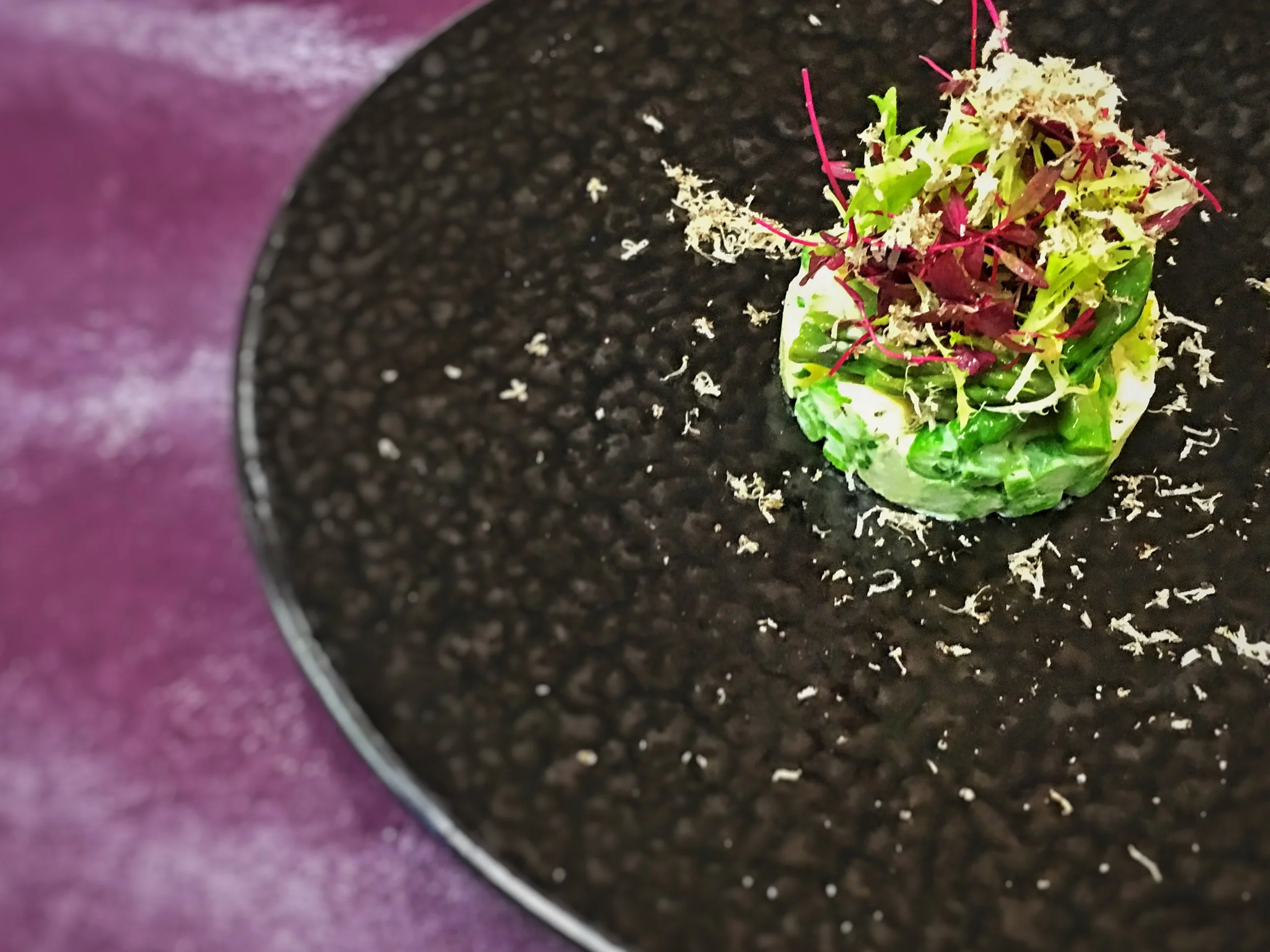 A small dish of microgreens and salad greens on a black textured plate with a purple background.
