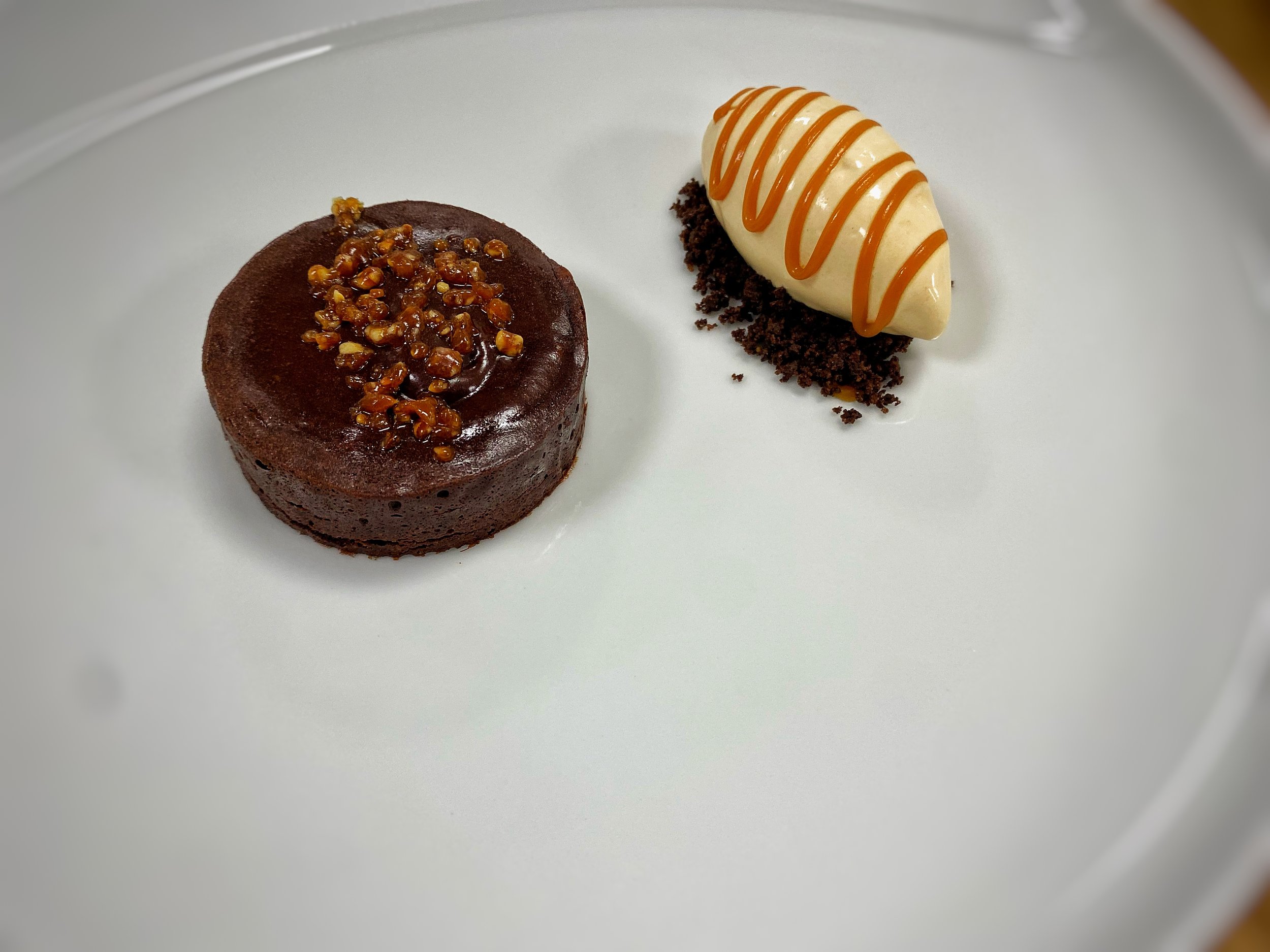 A white plate with two desserts: a round chocolate cake topped with caramelized nuts, and a scoop of vanilla ice cream drizzled with caramel sauce on a bed of crumbled brownie bits.