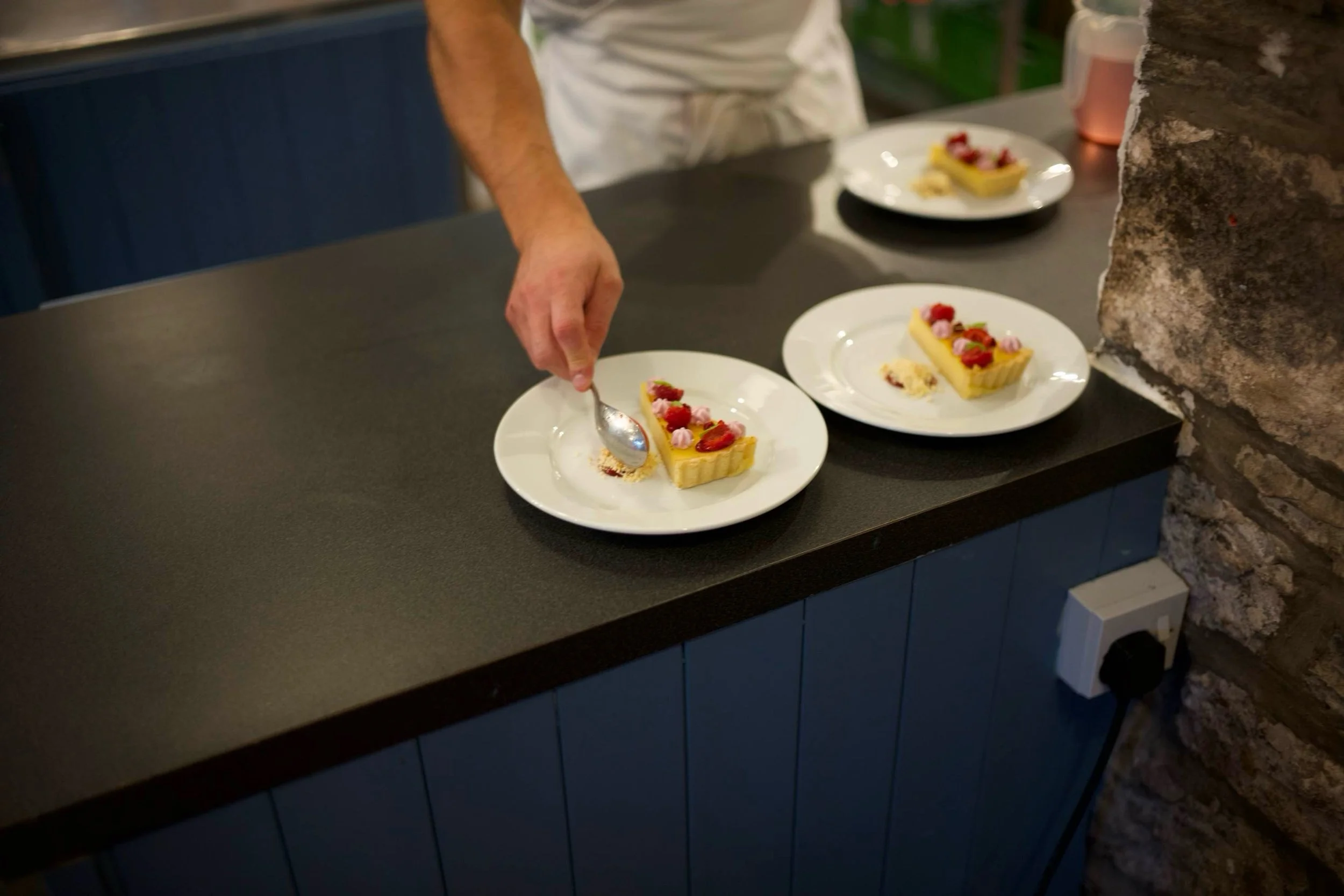 Person placing a dessert on a white plate with a spoon, with similar desserts on other plates on a dark countertop in a kitchen or restaurant setting.