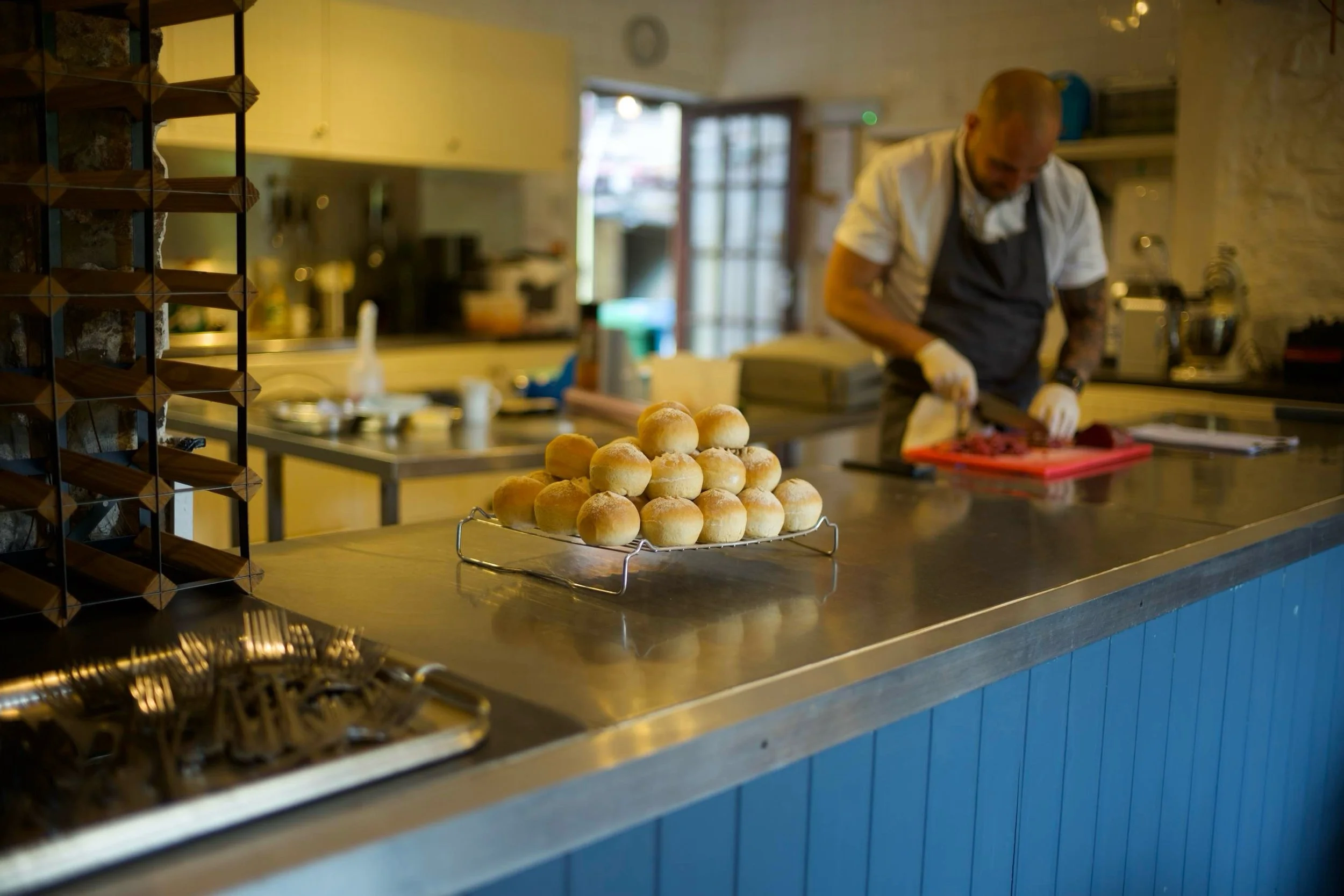 A worker slicing meat in a kitchen with a tray of baked bread rolls on the counter in the foreground.