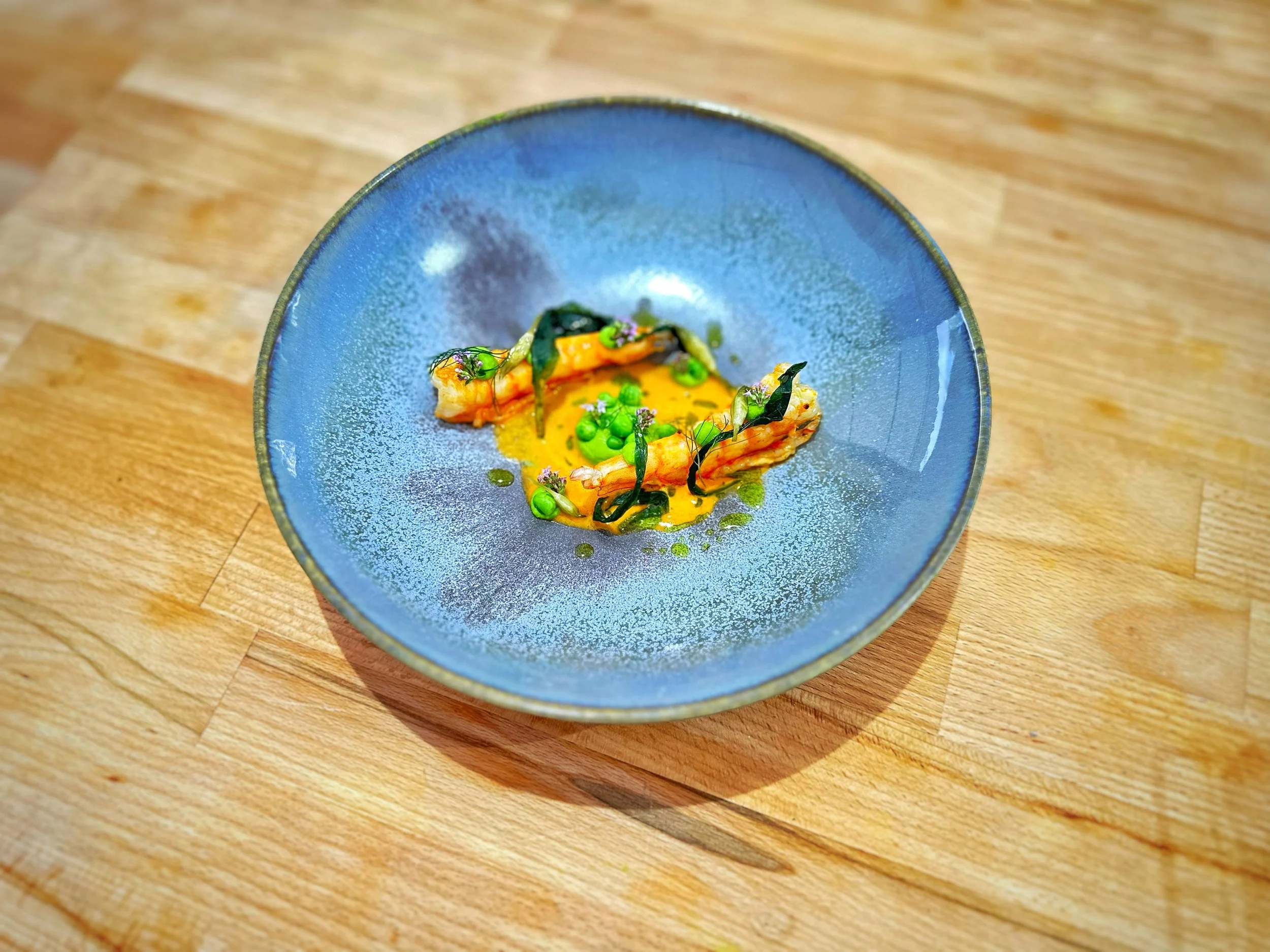 A blue ceramic plate with a small gourmet seafood dish garnished with green herbs and sauce, on a wooden surface.