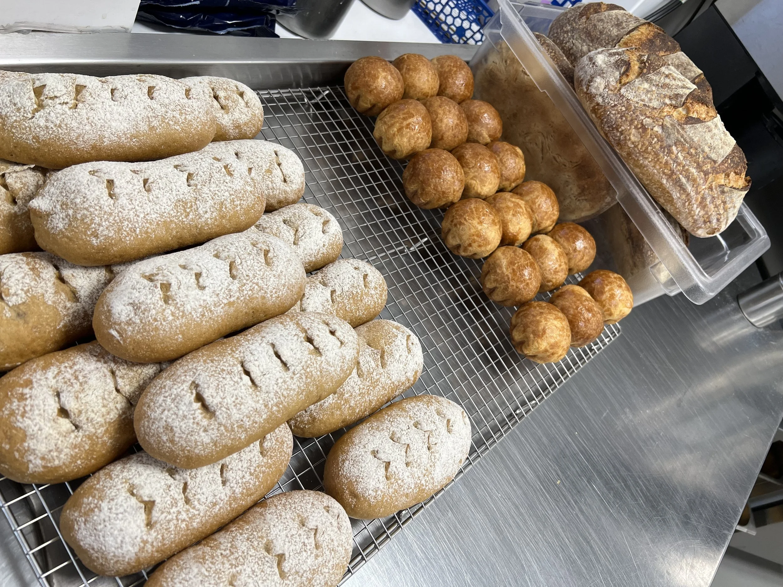 Assorted baked goods including eclairs dusted with powdered sugar, round bread rolls, and baguette on a metal cooling rack and clear plastic container.