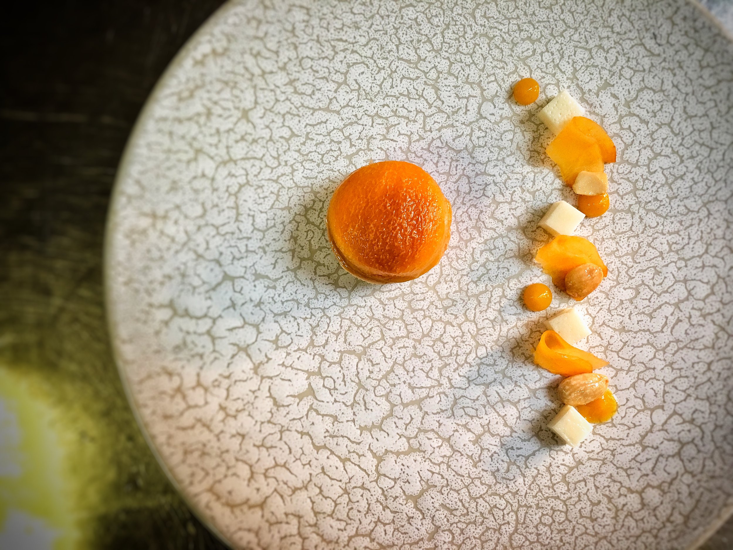 A white textured plate with a small round orange dessert or fruit and a curved line of small white, orange, and beige food garnishes