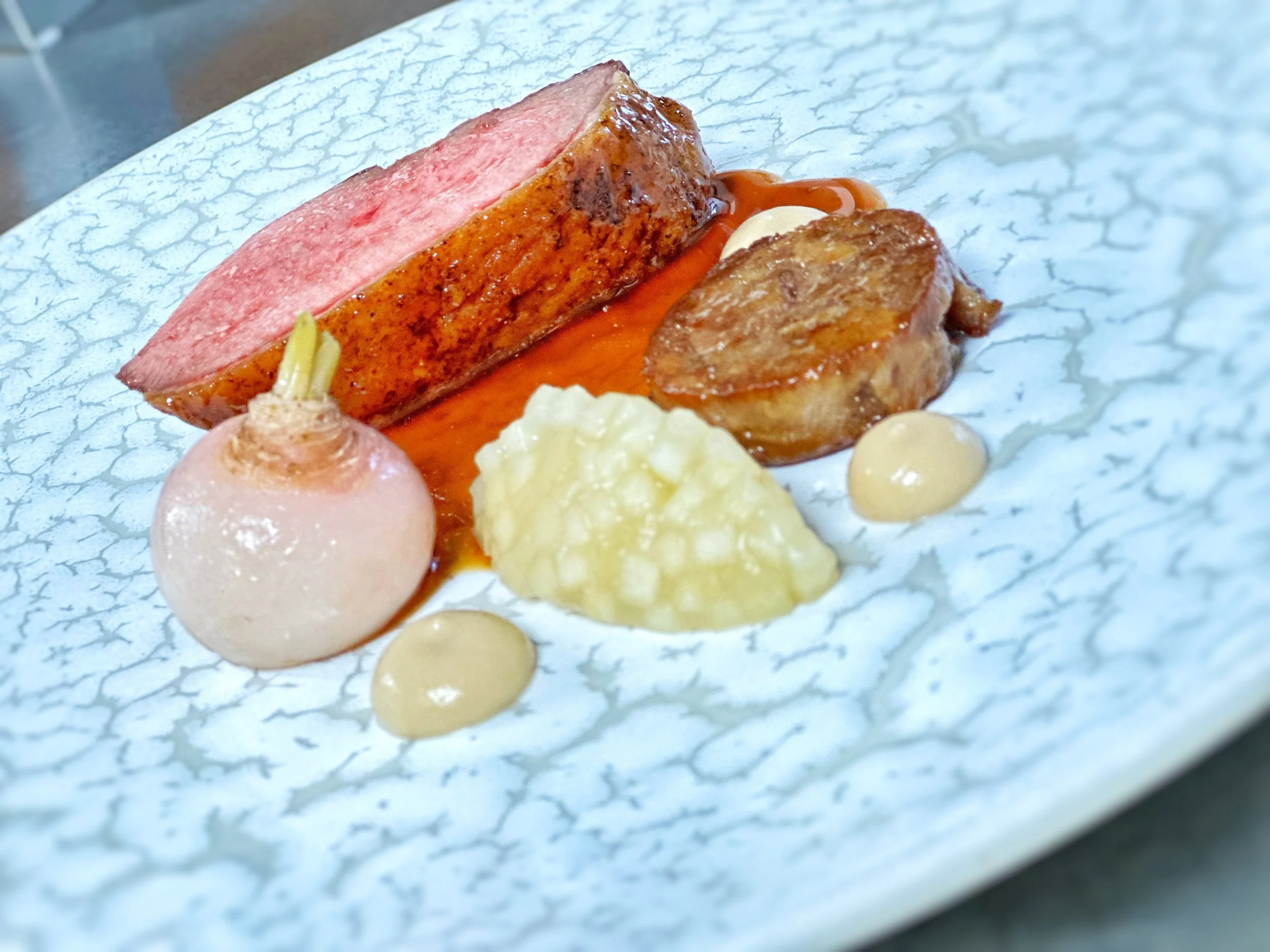 A plated gourmet dish featuring a thick slice of seared duck breast, a poached turnip, a confit leg in a cylinder a apple compote and turnip puree, finished with a sauce on a white textured plate.