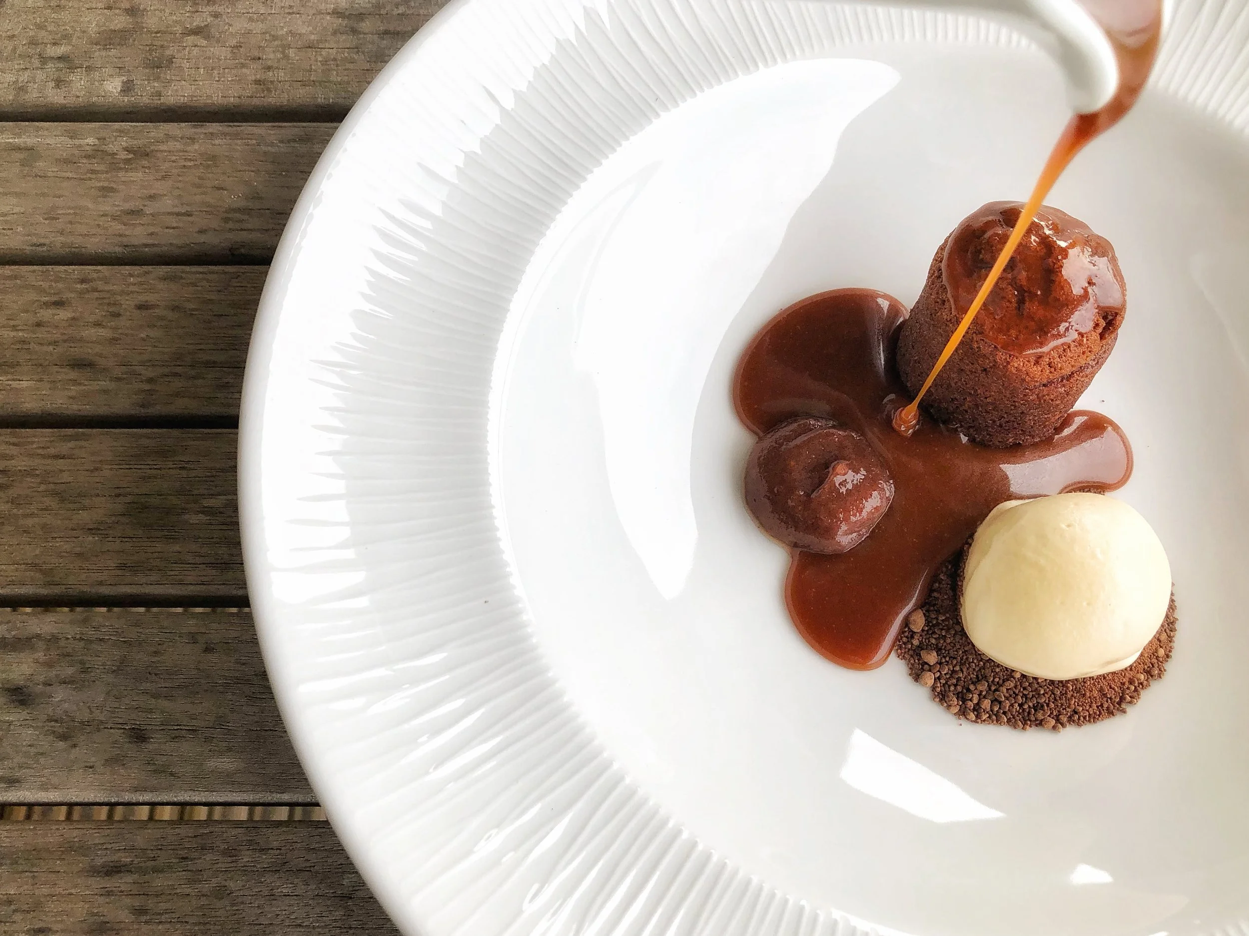 Chocolate lava cake with caramel sauce, served with a scoop of vanilla ice cream on a white plate, with a wooden table background.