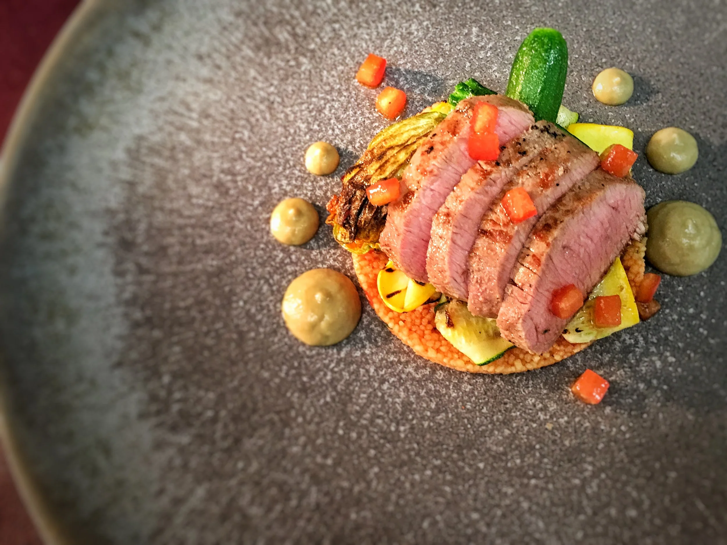 Slices of cooked steak served on a bed of roasted vegetables with dollops of sauce around the dish.