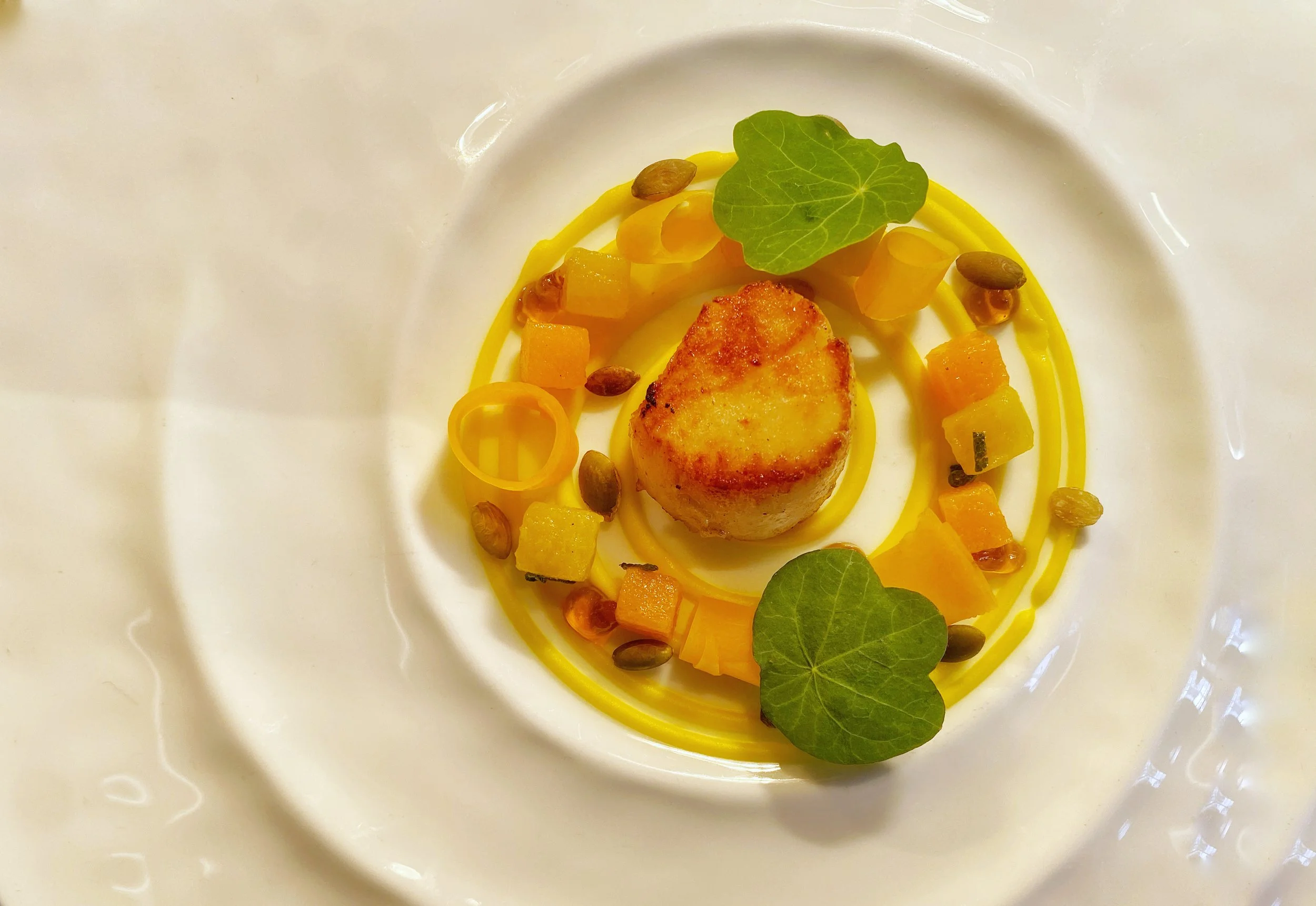A gourmet dish featuring a seared scallop centered on a white plate, garnished with small yellow and orange diced vegetables, pumpkin seeds, a spiral yellow vegetable or sauce, and garnished with green leaves.