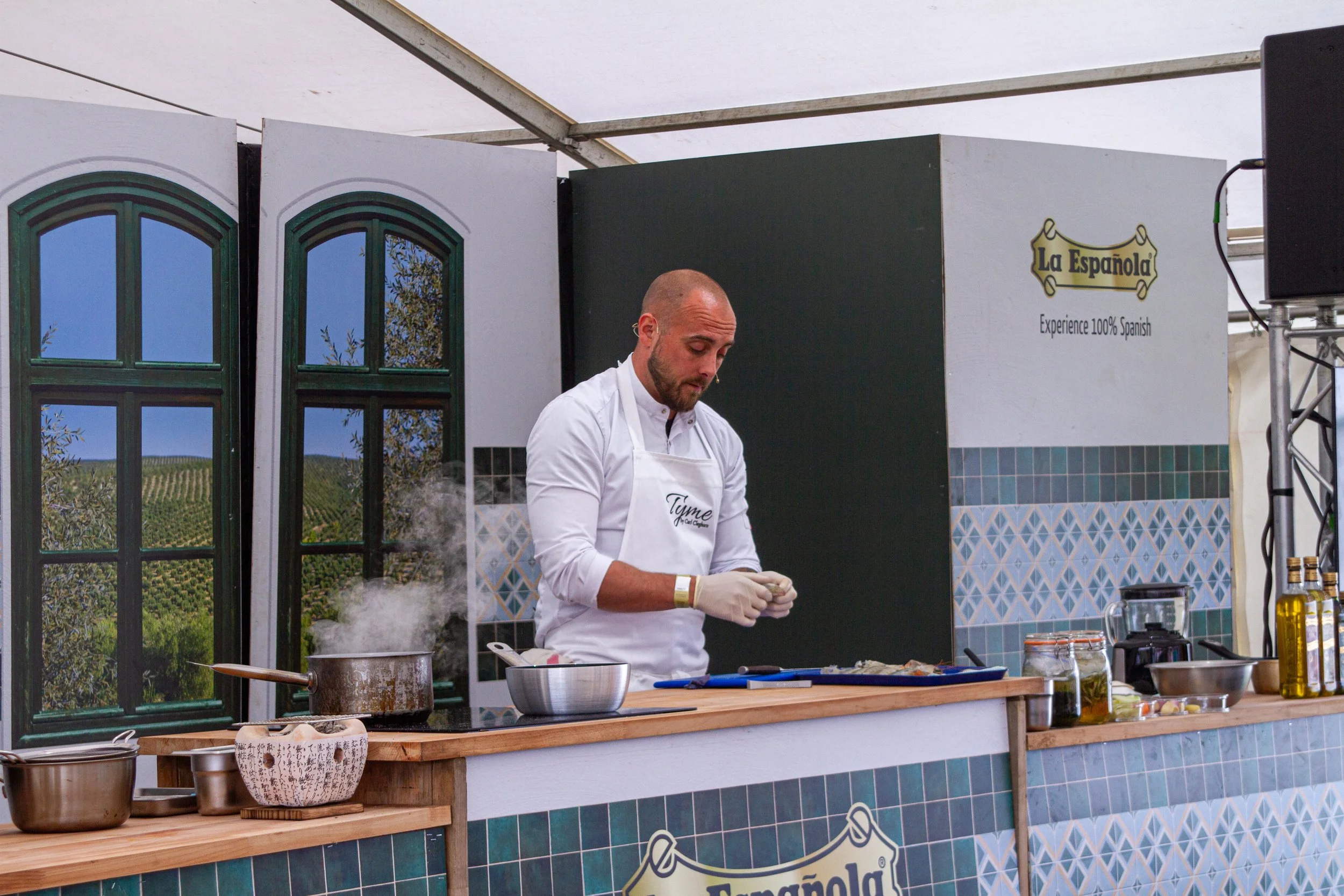 A chef Carl Cleghorn in a white apron preparing food on a wooden counter at an outdoor kitchen setup with scenic vineyard backdrop and decorative Spanish-style window panels.
