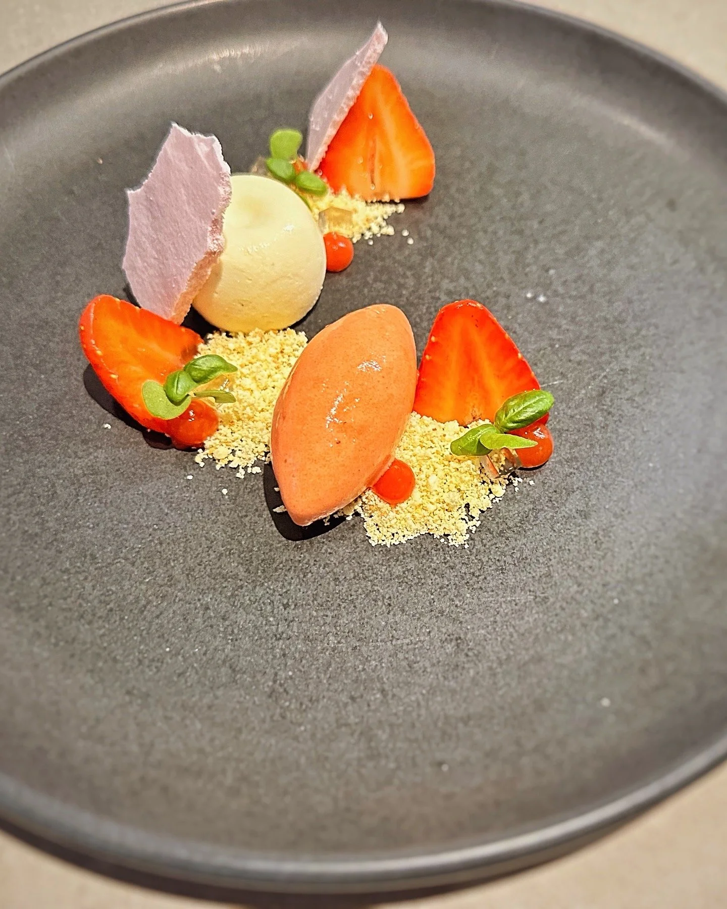 A dessert plate featuring strawberries, quenelle of white ice cream, orange-colored sorbet, chocolate shards, microgreens, and crumble or dusting on a dark round plate.