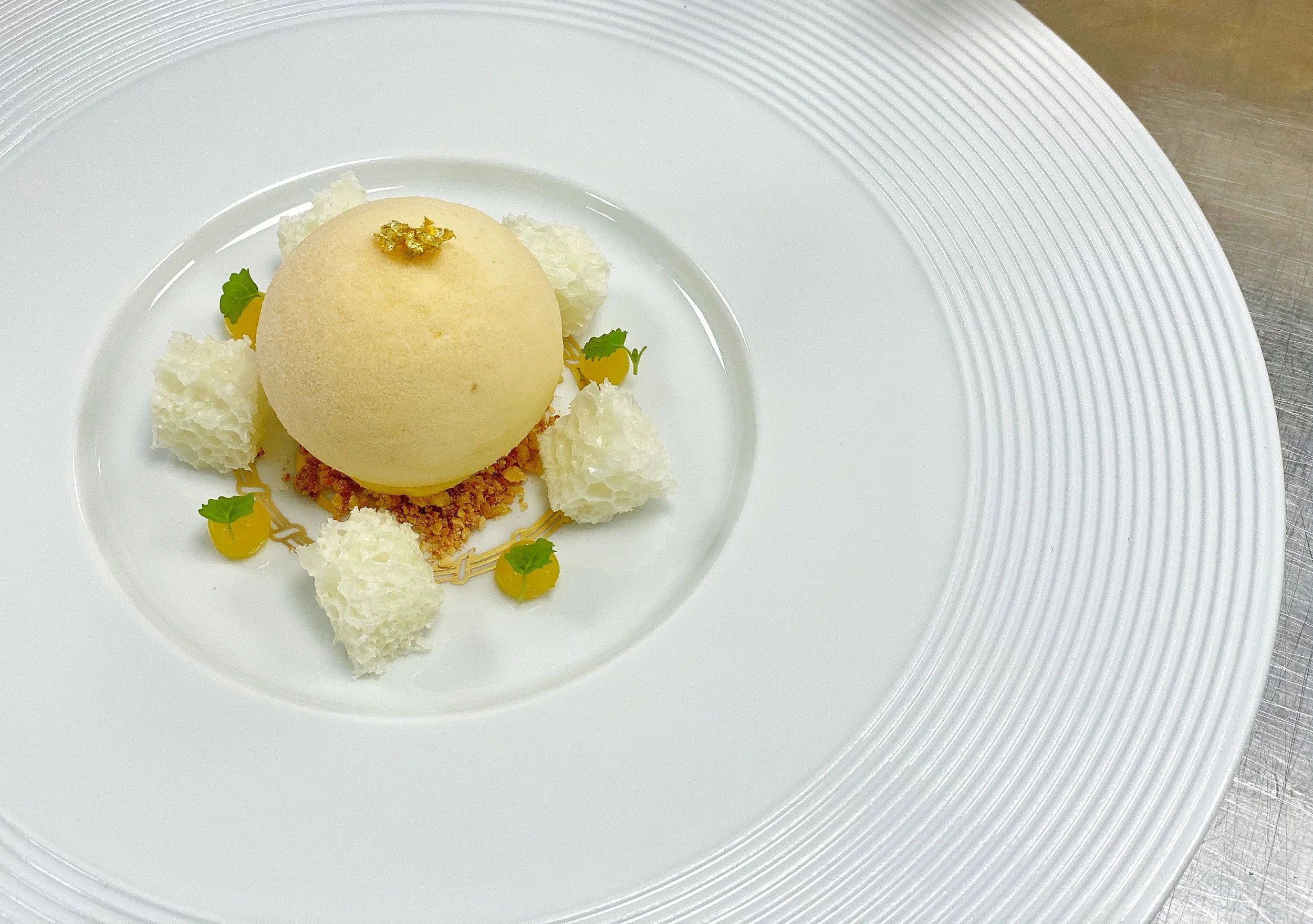 A plated dessert with a yellow sorbet or mousse sphere, garnished with a small amount of gold leaf on top, surrounded by white crispy elements, small green leaves, and yellow sauces on a white, textured-rim plate.