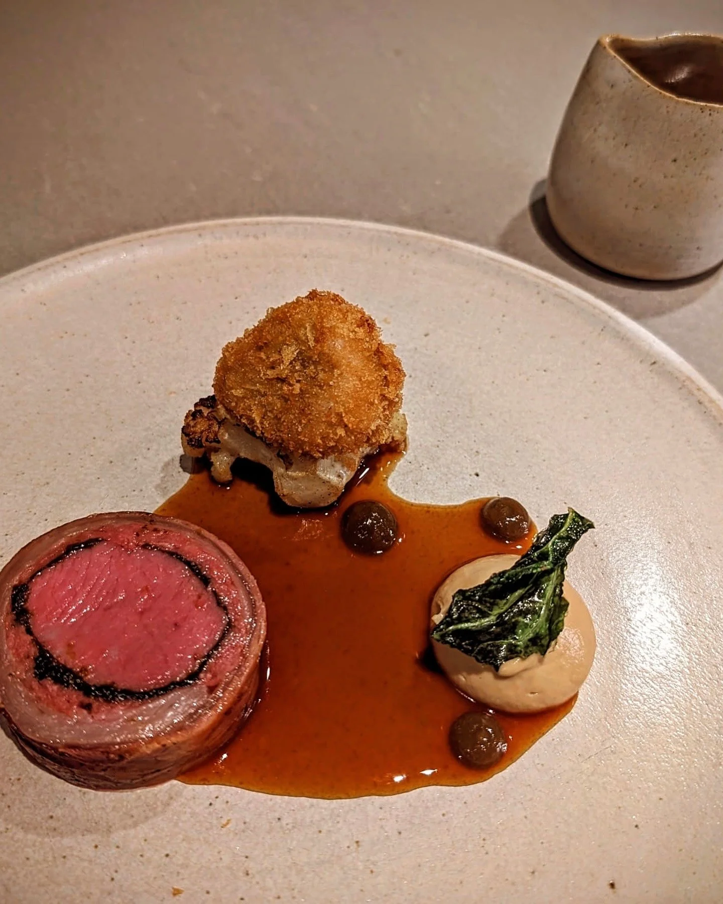 Elegant plated dish featuring a rolled piece of meat, a dollop of sauce with a green garnish, a piece of roasted or fried vegetable topped with a sauce, and small round black elements, all on a white plate with a small cream-colored pitcher in the ba