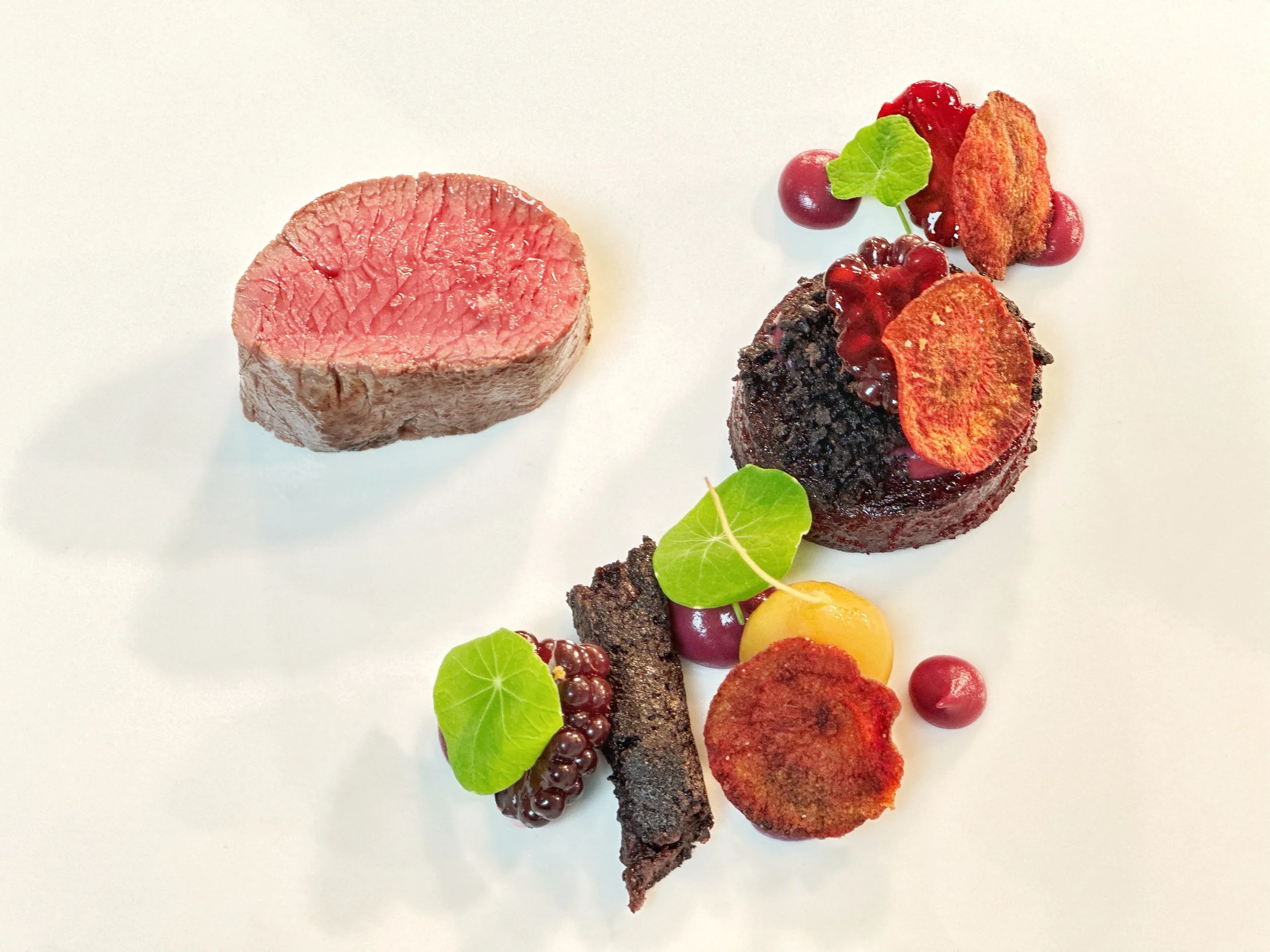 Seared medium-rare venison, garnished with beetroot fondant, crispy black pudding, blackberry fruit and herbs, plated on a white background