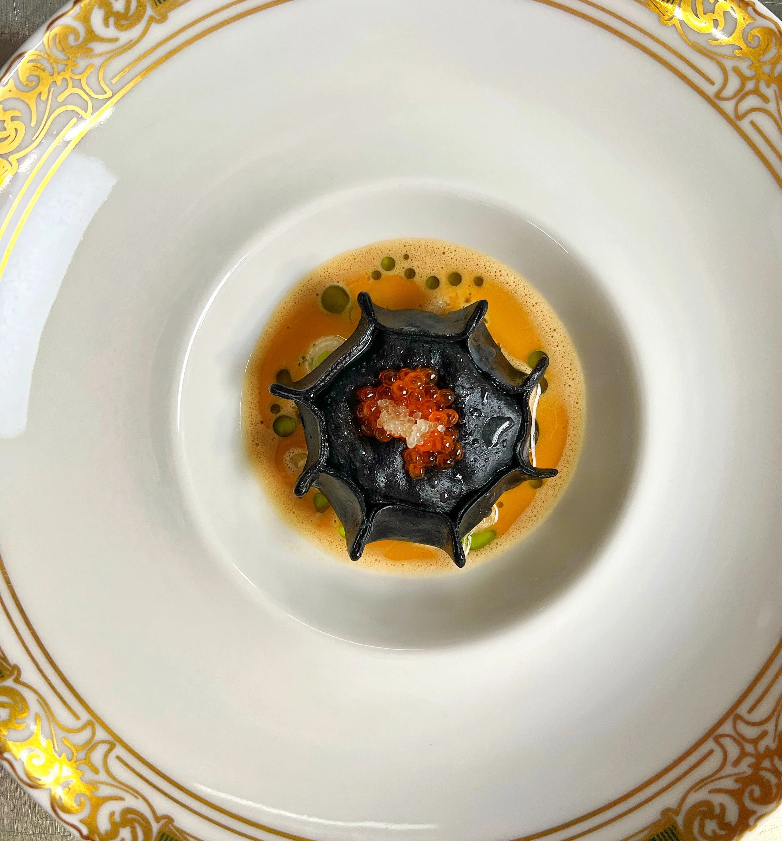 A dish featuring black pasta shaped like a spider web, topped with orange and white fish roe, served in a shallow bowl with ornate gold detailing on the rim. The bowl contains a yellow-orange broth or sauce with small green garnish.