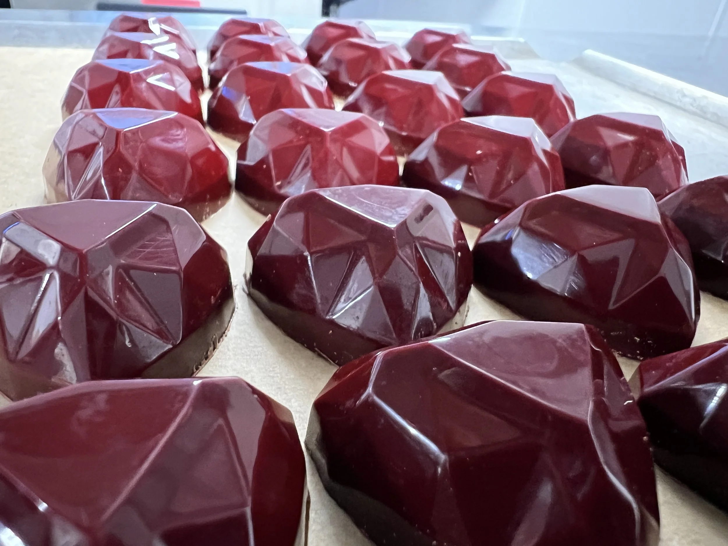 A close-up image of multiple red, faceted, geometric-shaped chocolates or desserts arranged on a surface.