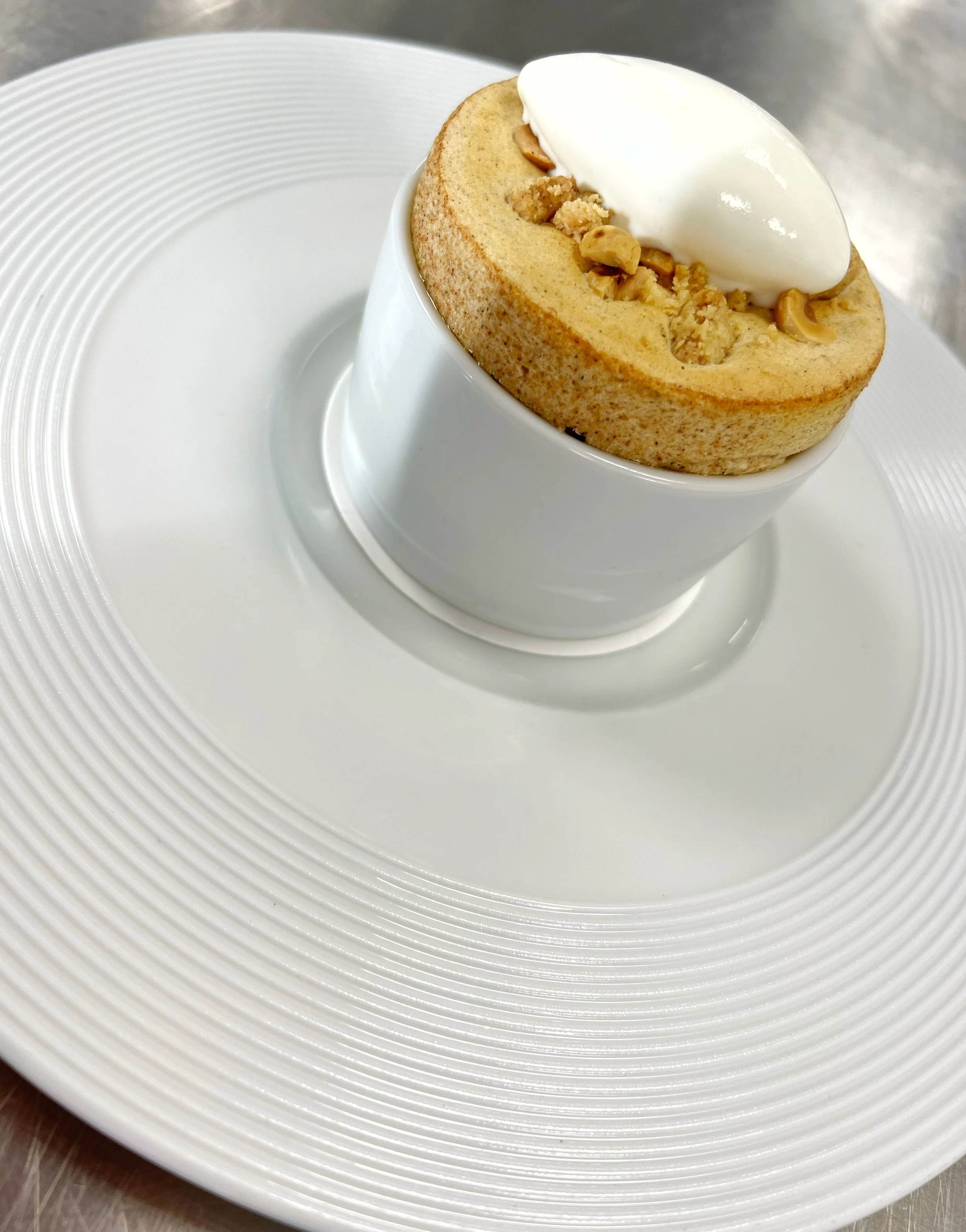 A scuffle topped  with nuts and a dollop of cream on top, served in a white ceramic cup on a white plate.