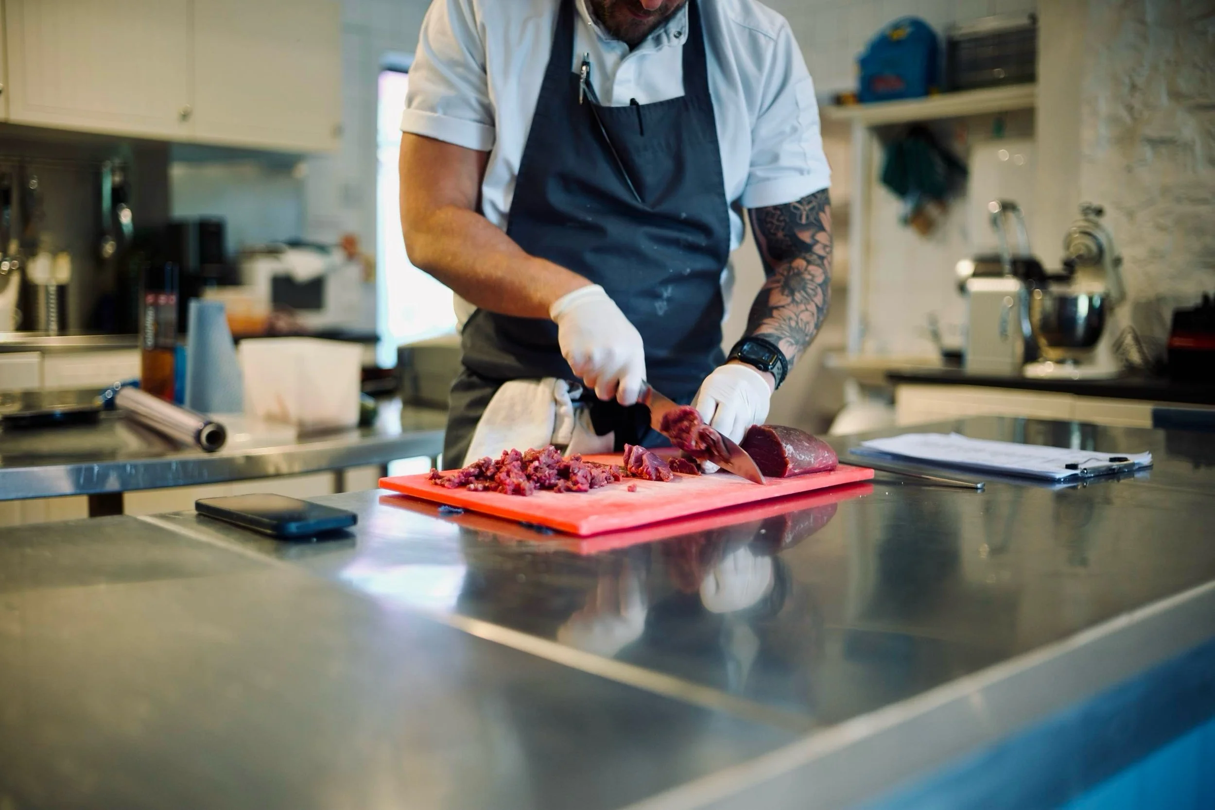 A chef in a white shirt and black apron is chopping raw beef on a red cutting board in a professional kitchen.