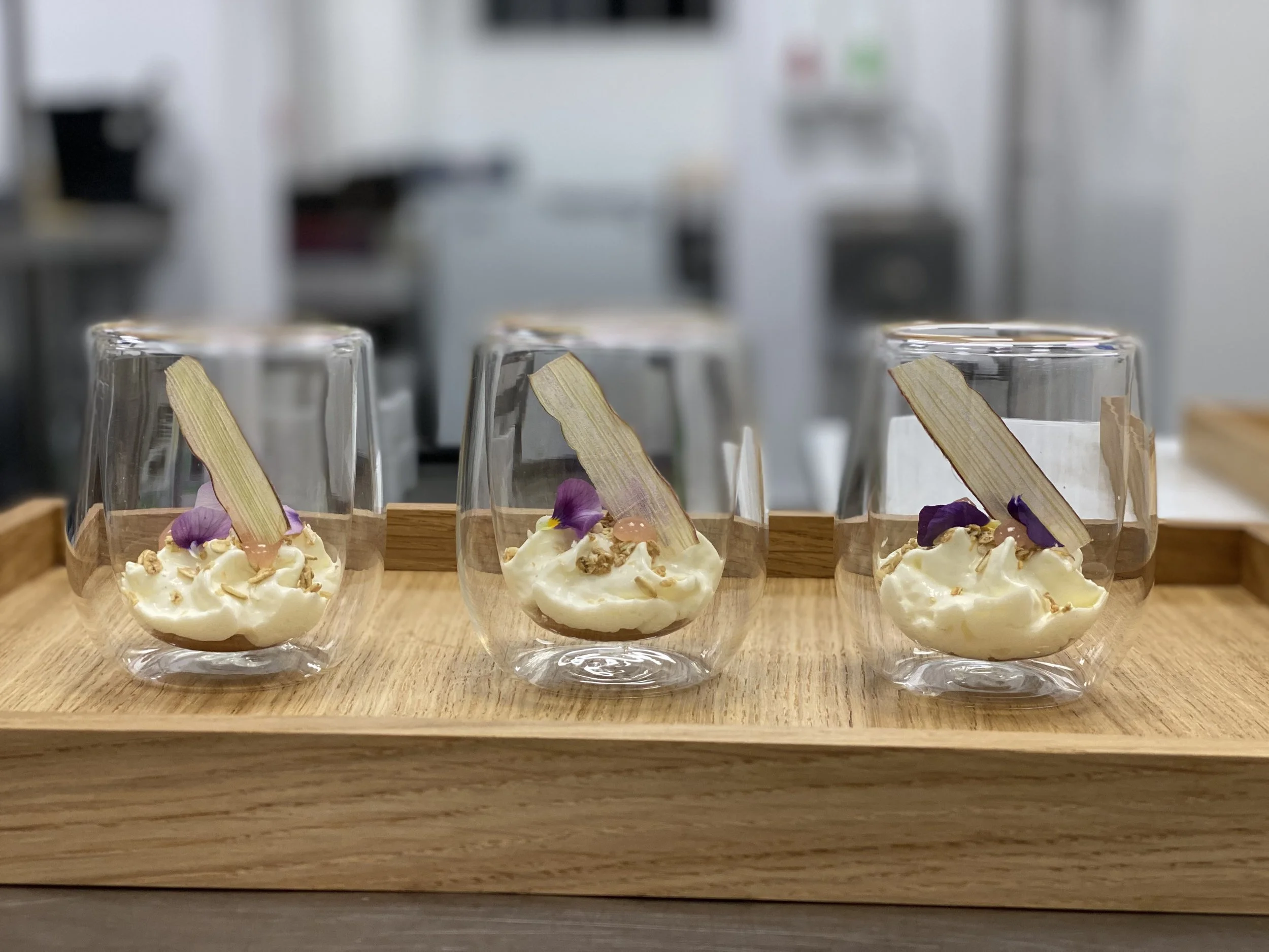 Three dessert glasses with cream, garnished with purple edible flowers, a thin rectangular chocolate, and some small crunchy toppings, placed on a wooden tray.