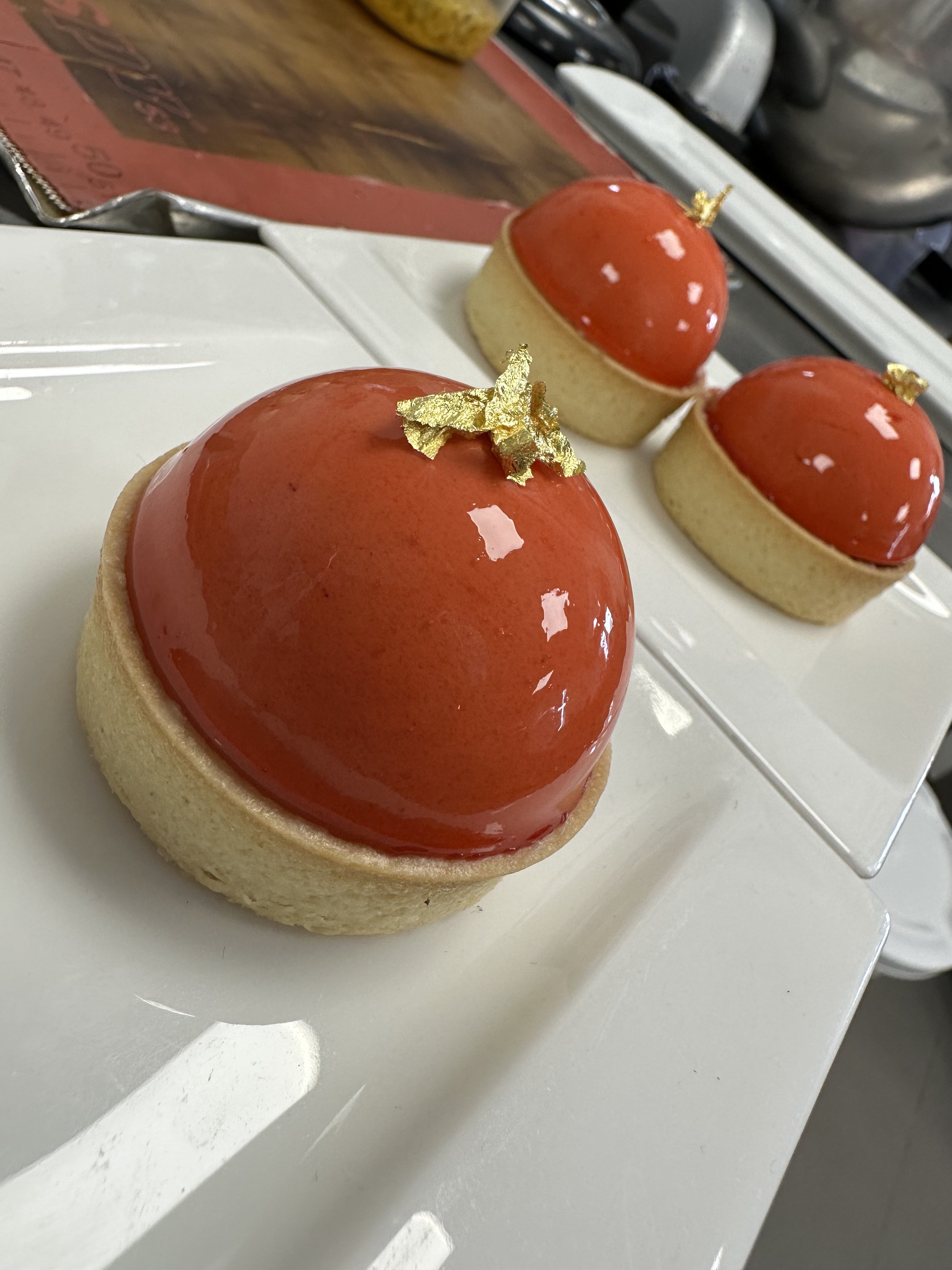 Three round pastries with shiny red glaze and thin golden leaf decorations, arranged on a white rectangular plate.