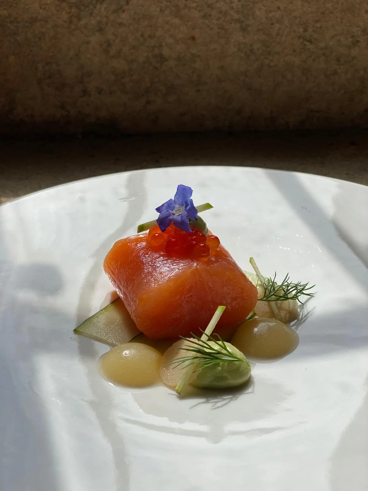 A plated dish featuring a piece of smoked salmon garnished with a small blue edible flower, red and yellow caviar, and accompanied by slices of cucumber and sprigs of dill on a white plate.