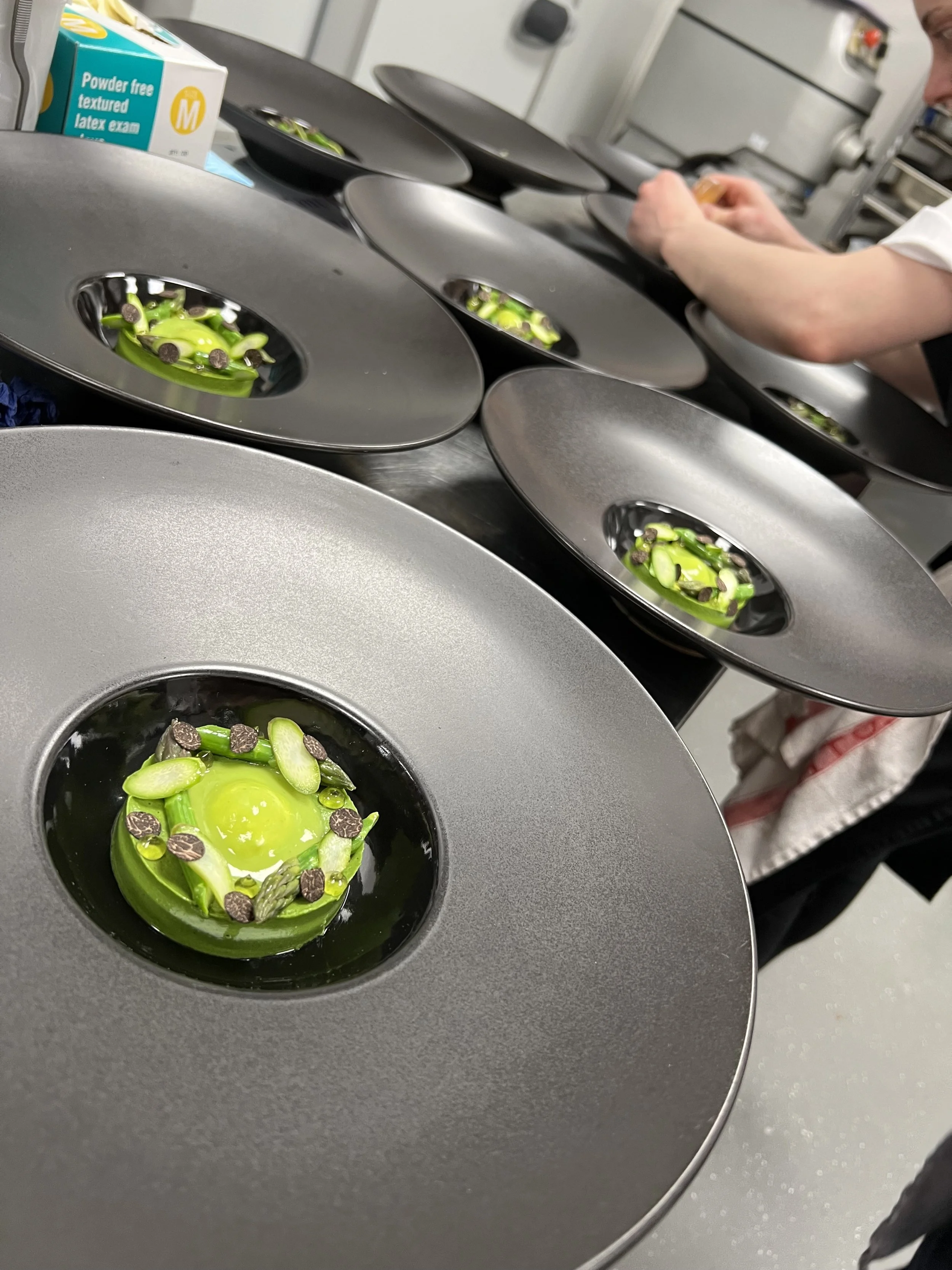 Several black plates with small, green, circular molded desserts topped with black seeds, possibly chocolate seeds, arranged in a circular pattern. In the background, a person is preparing or decorating more desserts in a kitchen setting.