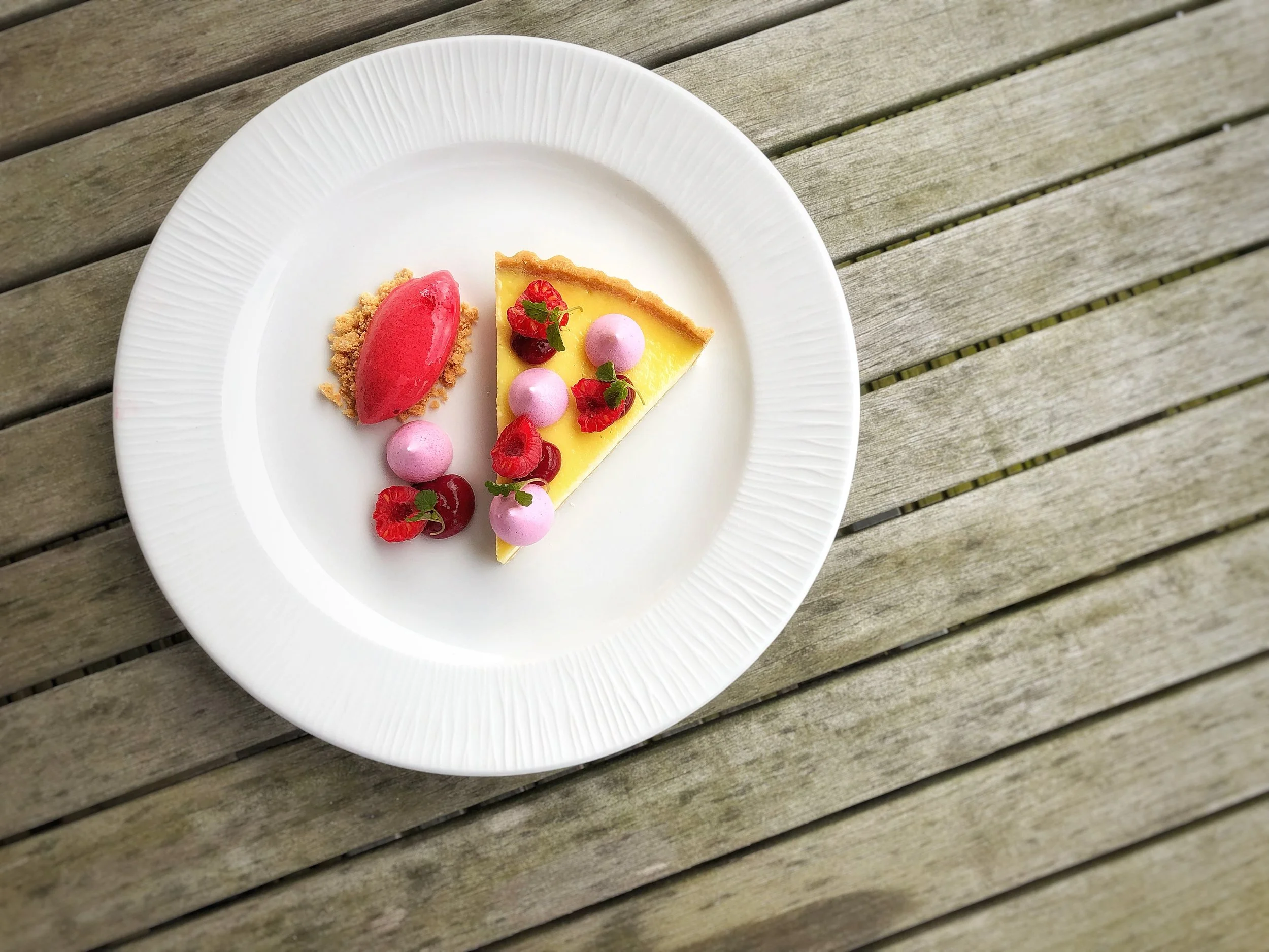A white plate with a slice of lemon tart decorated with pink meringue dollops, fresh raspberries, and edible flower petals, next to a scoop of pink sorbet with a crumbly base, all on a wooden surface.