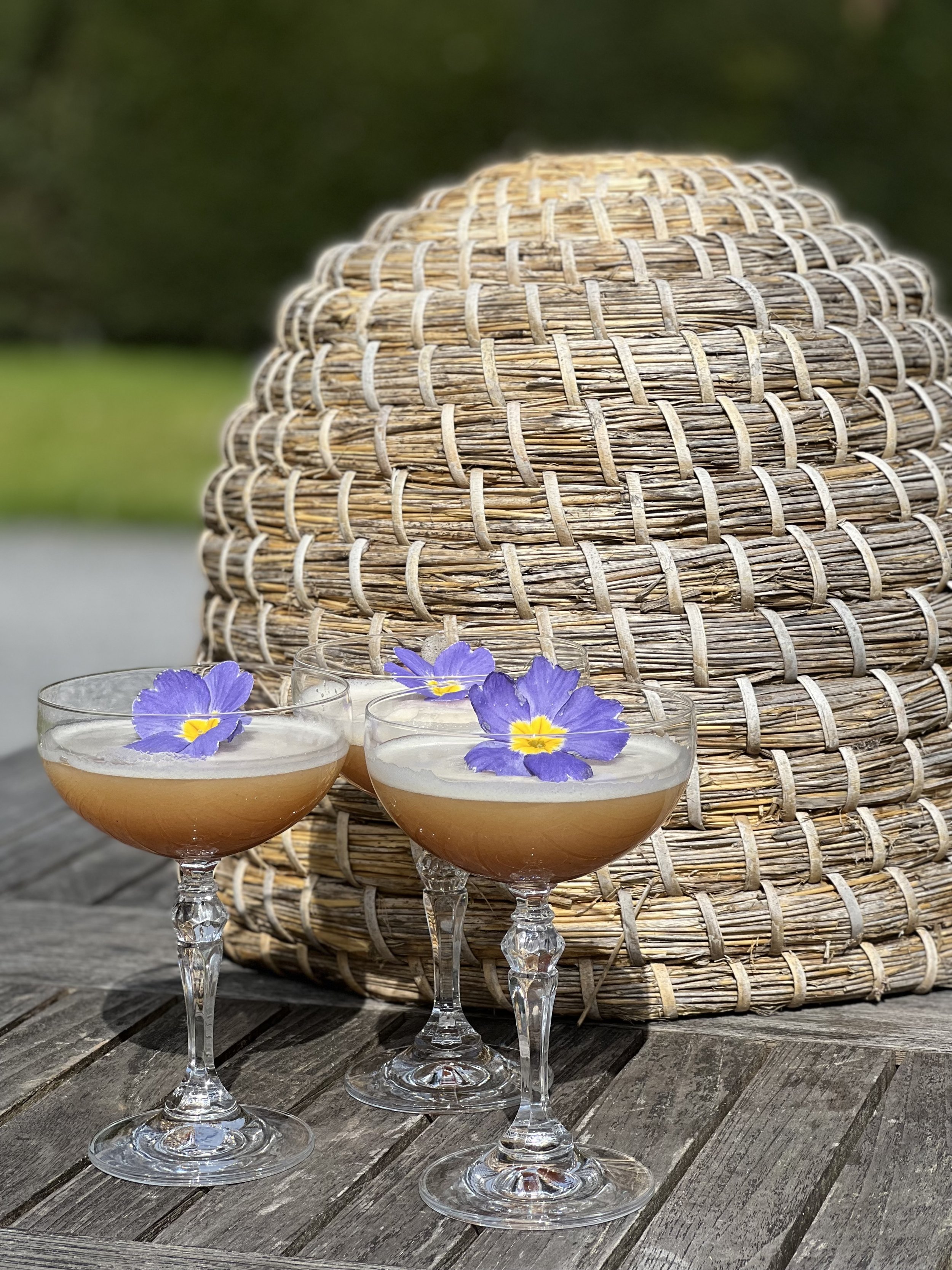 Three coupe glasses with pinkish drinks, garnished with purple flowers, placed outdoors on a wooden surface in front of a large woven straw hat or basket.