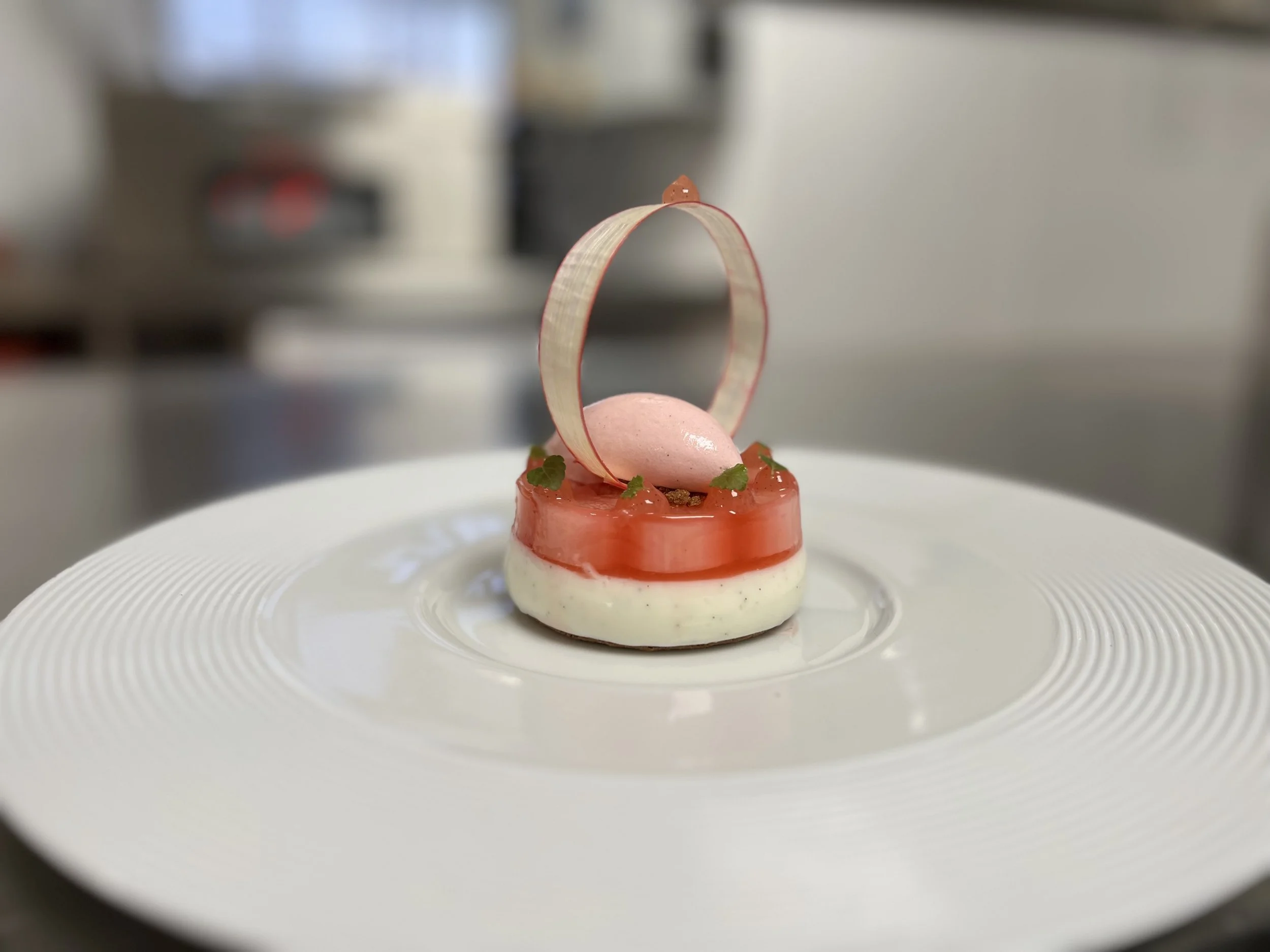 A deconstructed modern appetizer on a white textured plate, with a creamy white base, topped with a thin layer of red sauce or gel, decorated with small green herbs. There's a pink macaron and a delicate, curved sugar or candy strip as garnish.