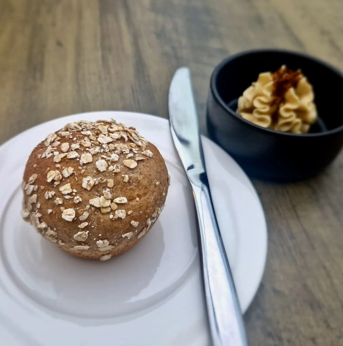 A muffin topped with oats on a white plate, a knife and spoon, with a bowl of butter and brown sugar next to it on a wooden table.