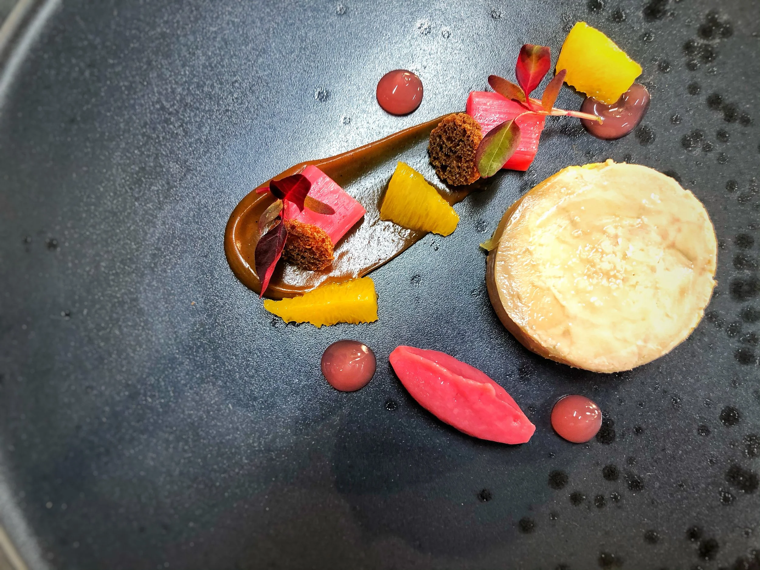 Elegant plated dessert with a round cheesecake, garnished with colorful fruit pieces, edible flowers, microgreens, and chocolate and fruit sauces on a black textured plate.