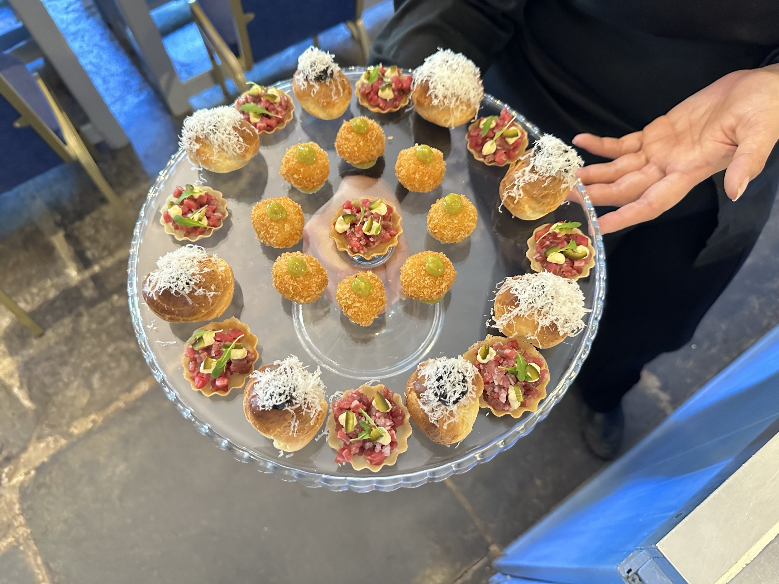A clear glass tray filled with assorted small, decorative appetizers and snacks, including pastry cups topped with chopped beef and greens, fried croquettes with green sauce, and mini cream puffs with shredded cheese or white topping.
