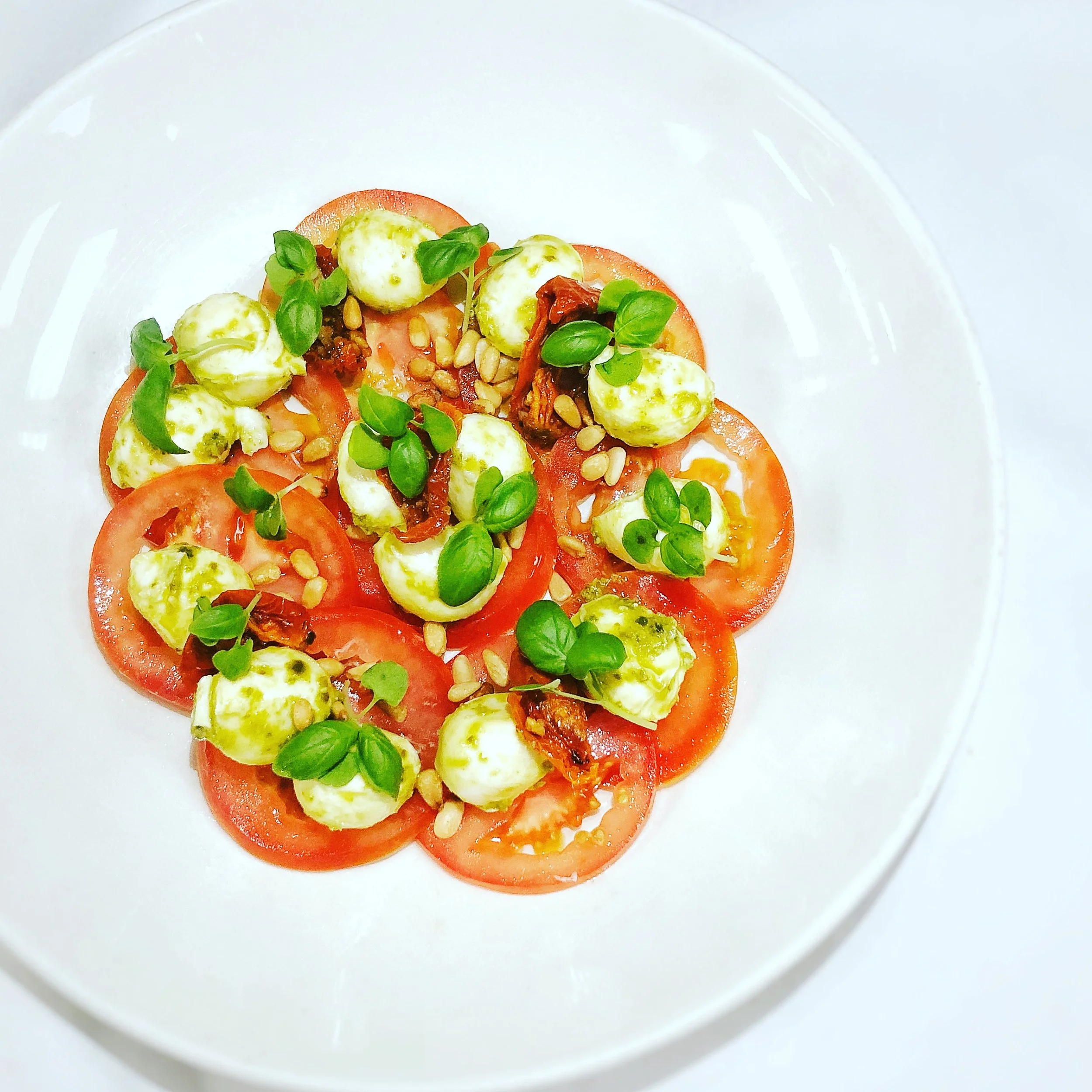 Tomato slices topped with mozzarella balls, fresh basil leaves, pine nuts, and sun-dried tomatoes on a white plate.