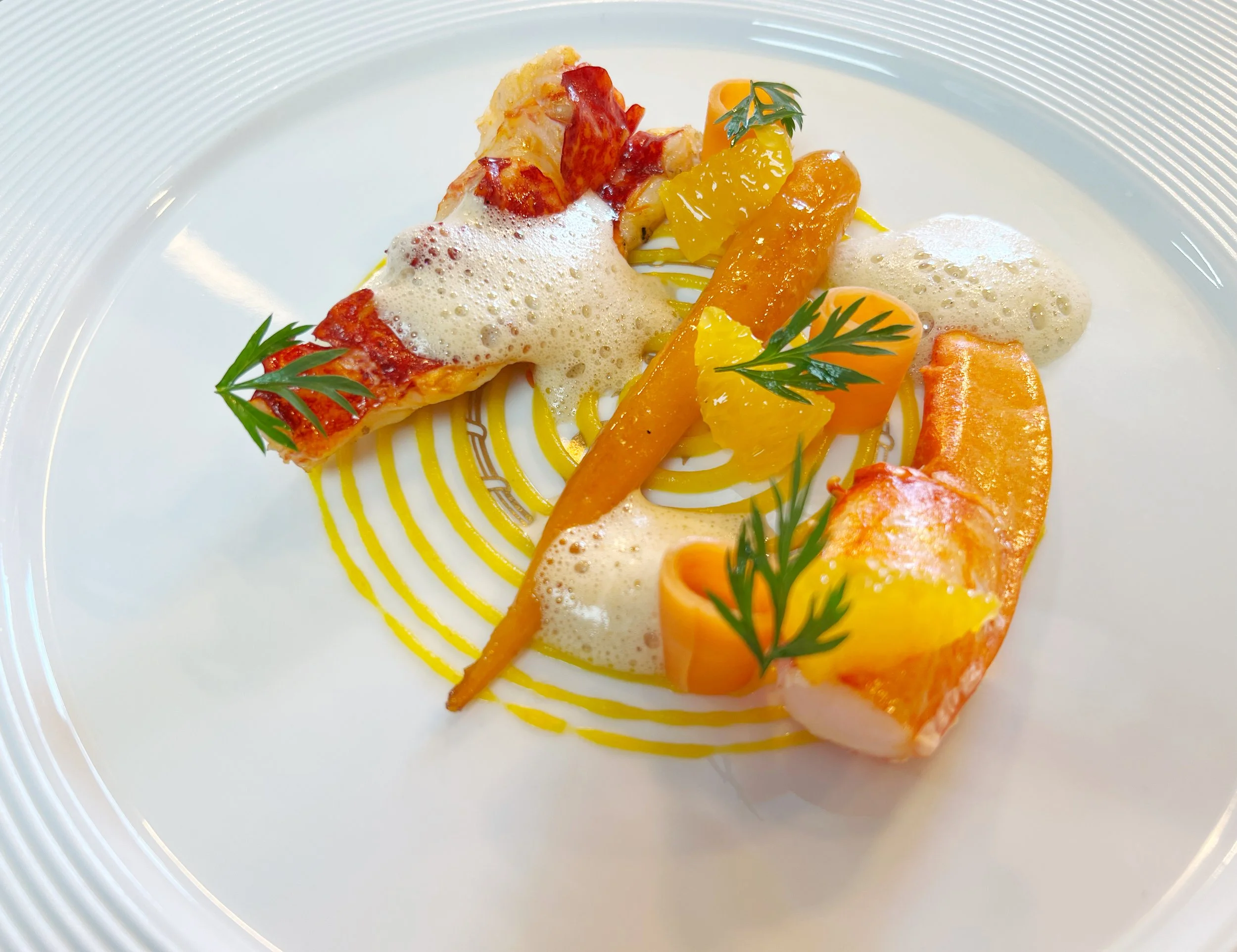 A plated gourmet dish featuring lobster with foam, yellow sauce, carrot, orange, and garnished with sprigs of fresh herbs, arranged artfully on a white plate.