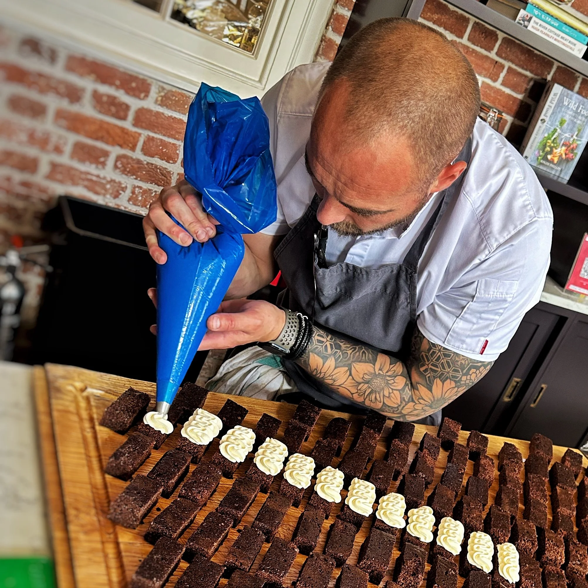 A man with tattoos on his arm decorating rectangular chocolate cakes with white frosting using a piping bag. He is leaning over a wooden board filled with rows of these cakes, in a kitchen with brick walls and bookshelves.