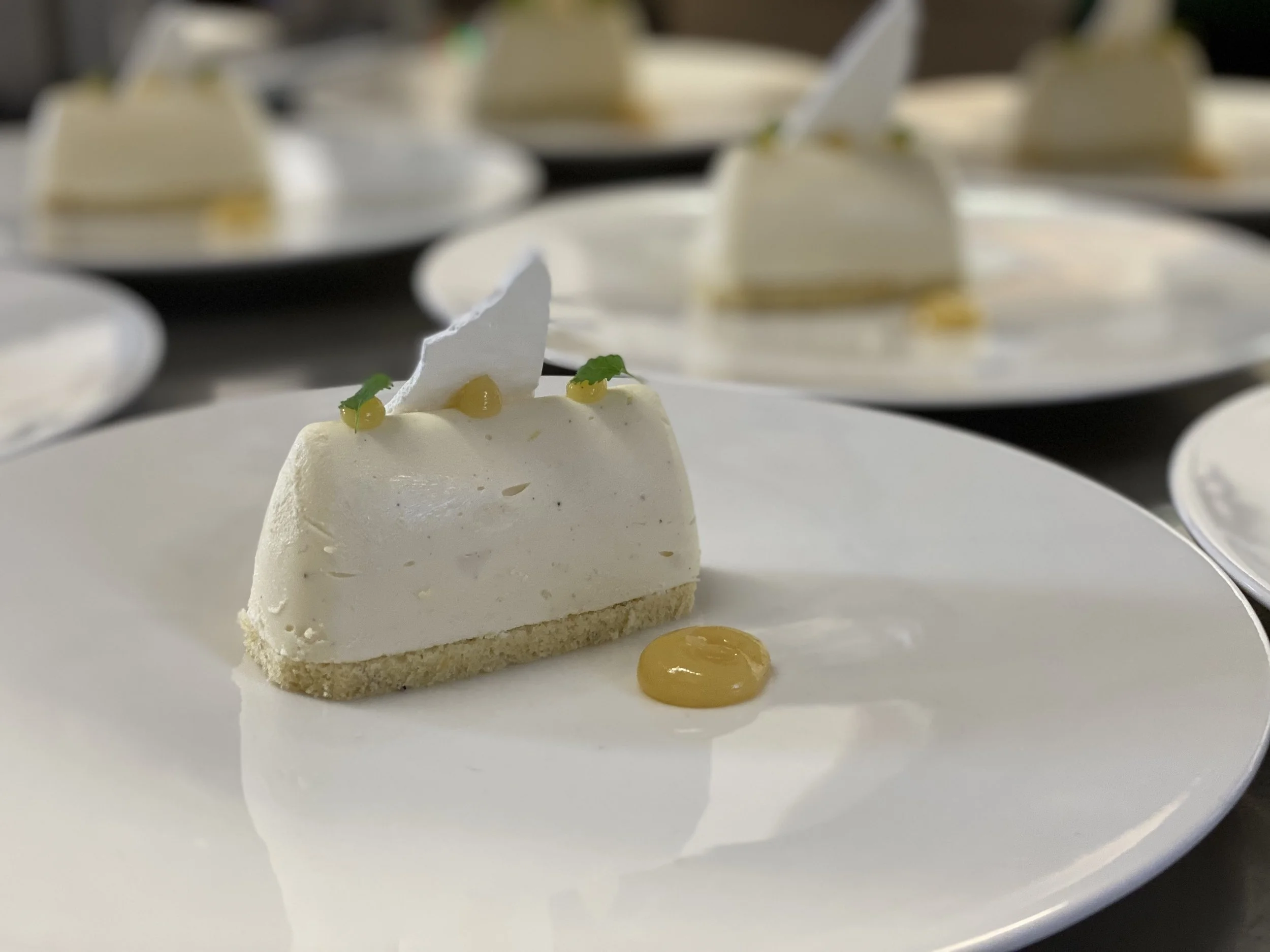 A slice of white cheesecake on a white plate, garnished with small green leaves, topped with a white piece of chocolate and yellow sauce dots, with more cheesecake slices on other plates in the background.