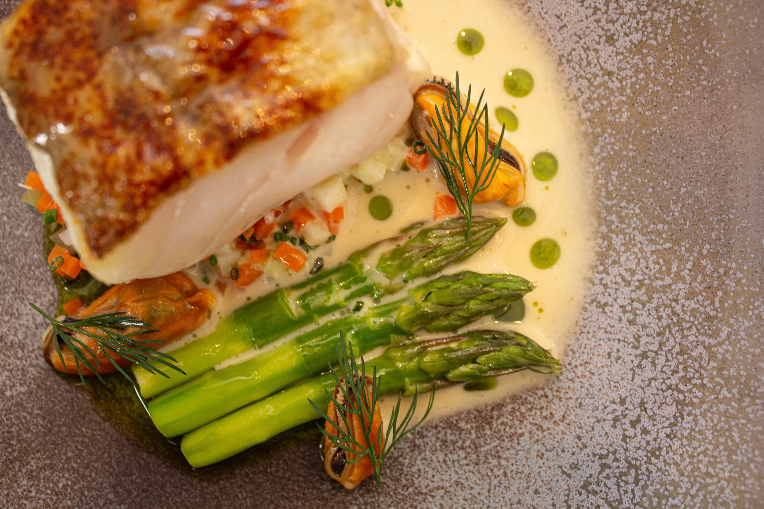 Pan-seared fish fillet served with grilled asparagus, mussels, chopped vegetables, and a creamy sauce garnished with dill and dotted with green oil droplets.