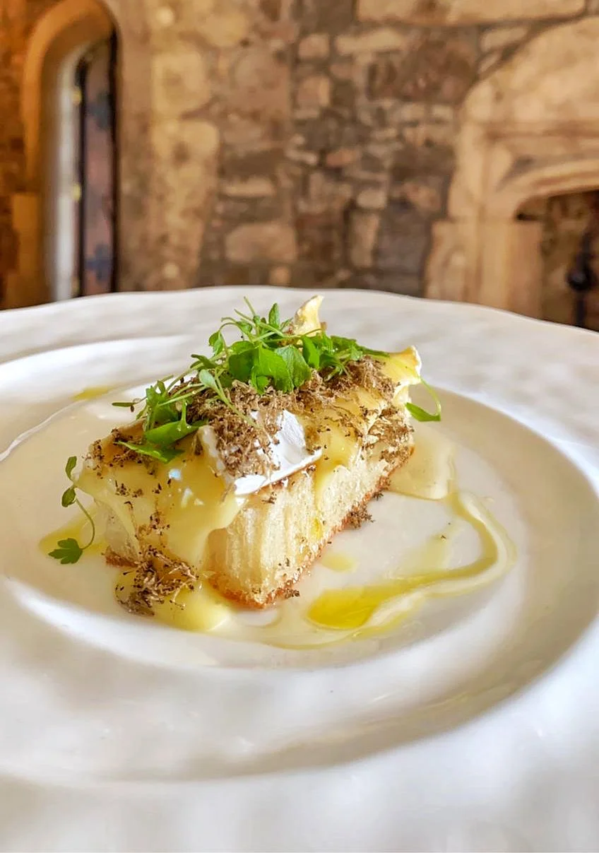 A slice of cheesecake topped with microgreens and drizzled with white chocolate and honey, served on a white plate with a wooden rustic background.