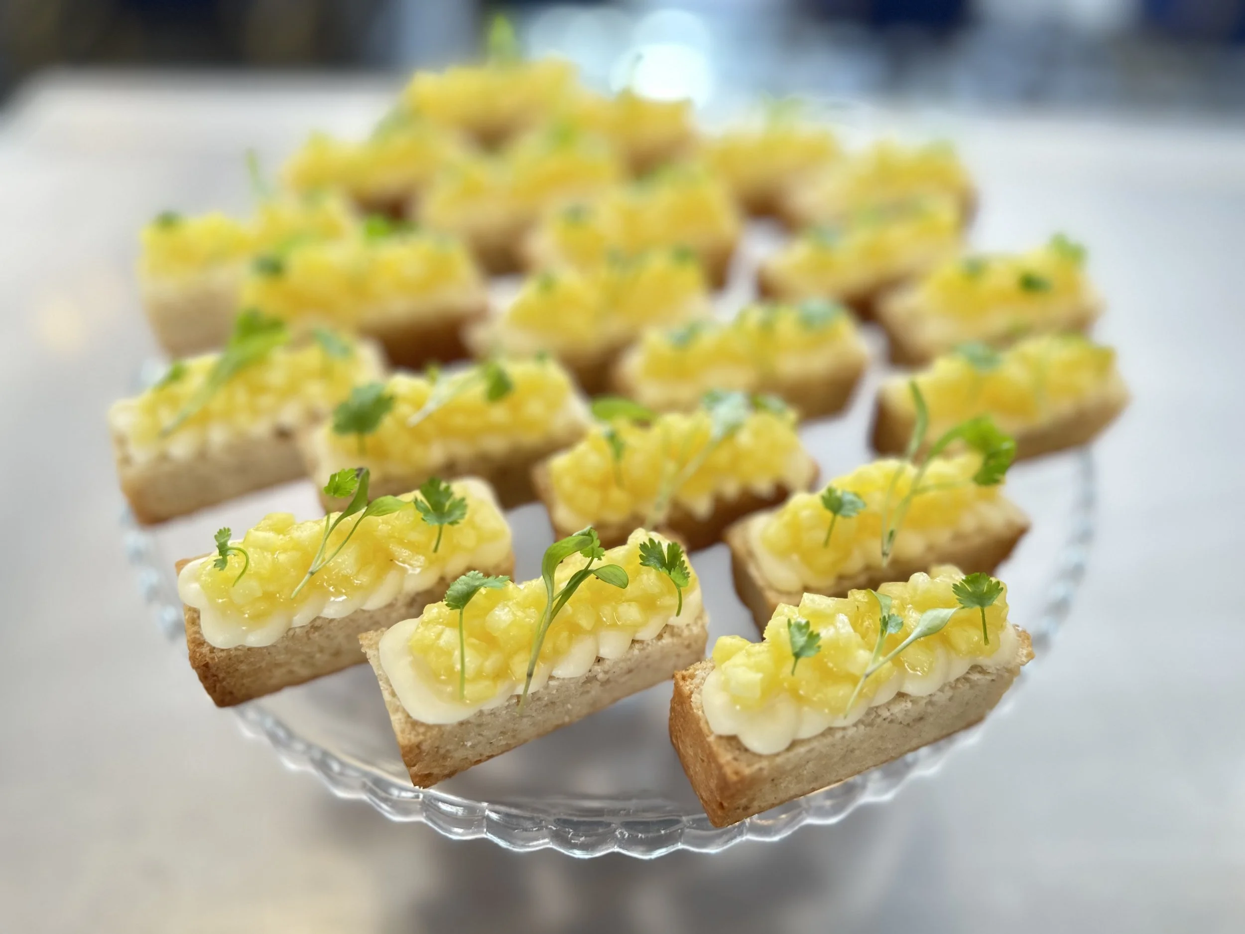 Tray of small toasted bread slices topped with yellowish spread, garnished with green herbs, arranged on a glass platter.