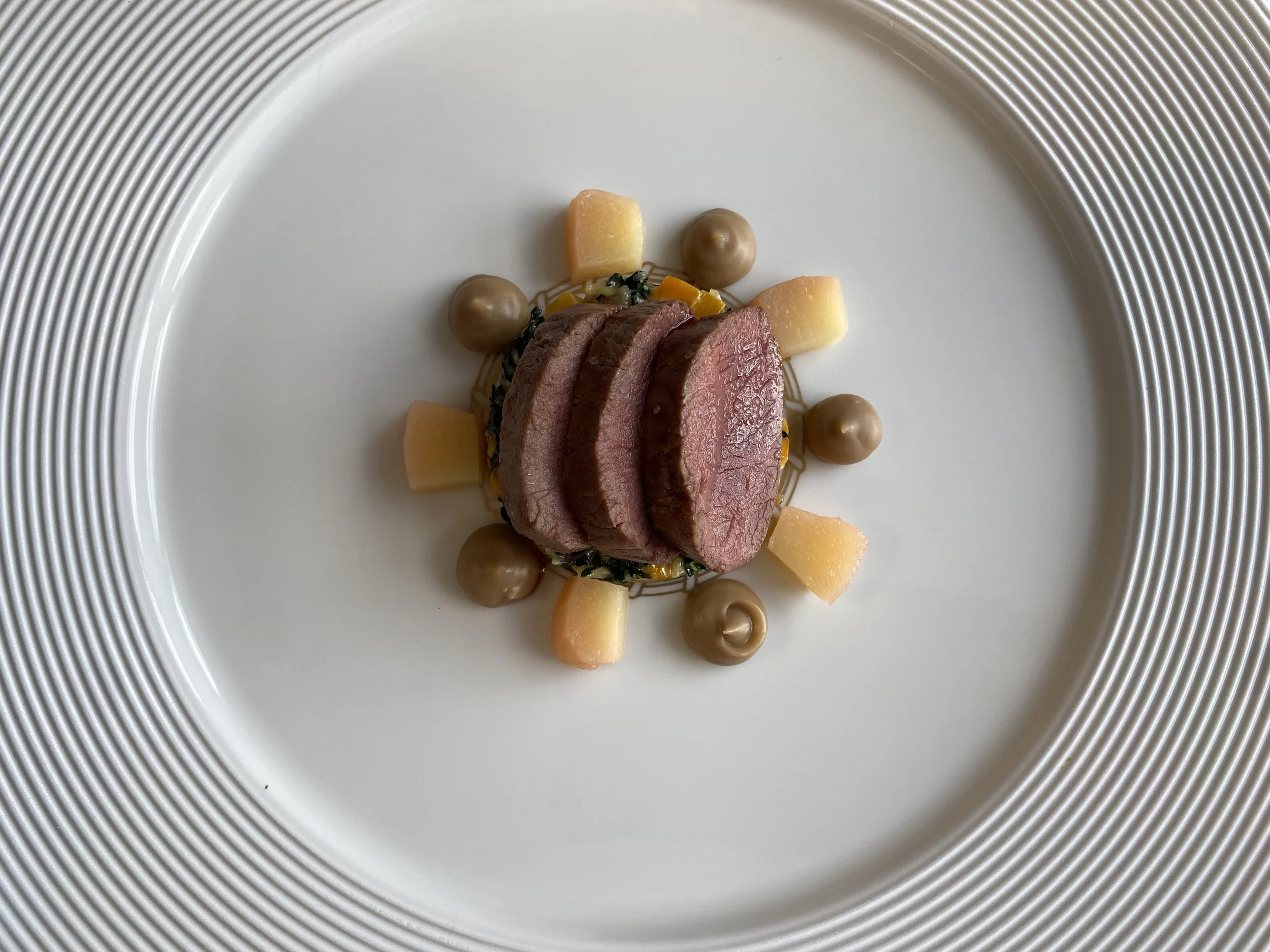 Slices of cooked meat arranged on a white plate with decorative elements including small dollops of brown sauce, yellow cubed vegetables, and green sauce.