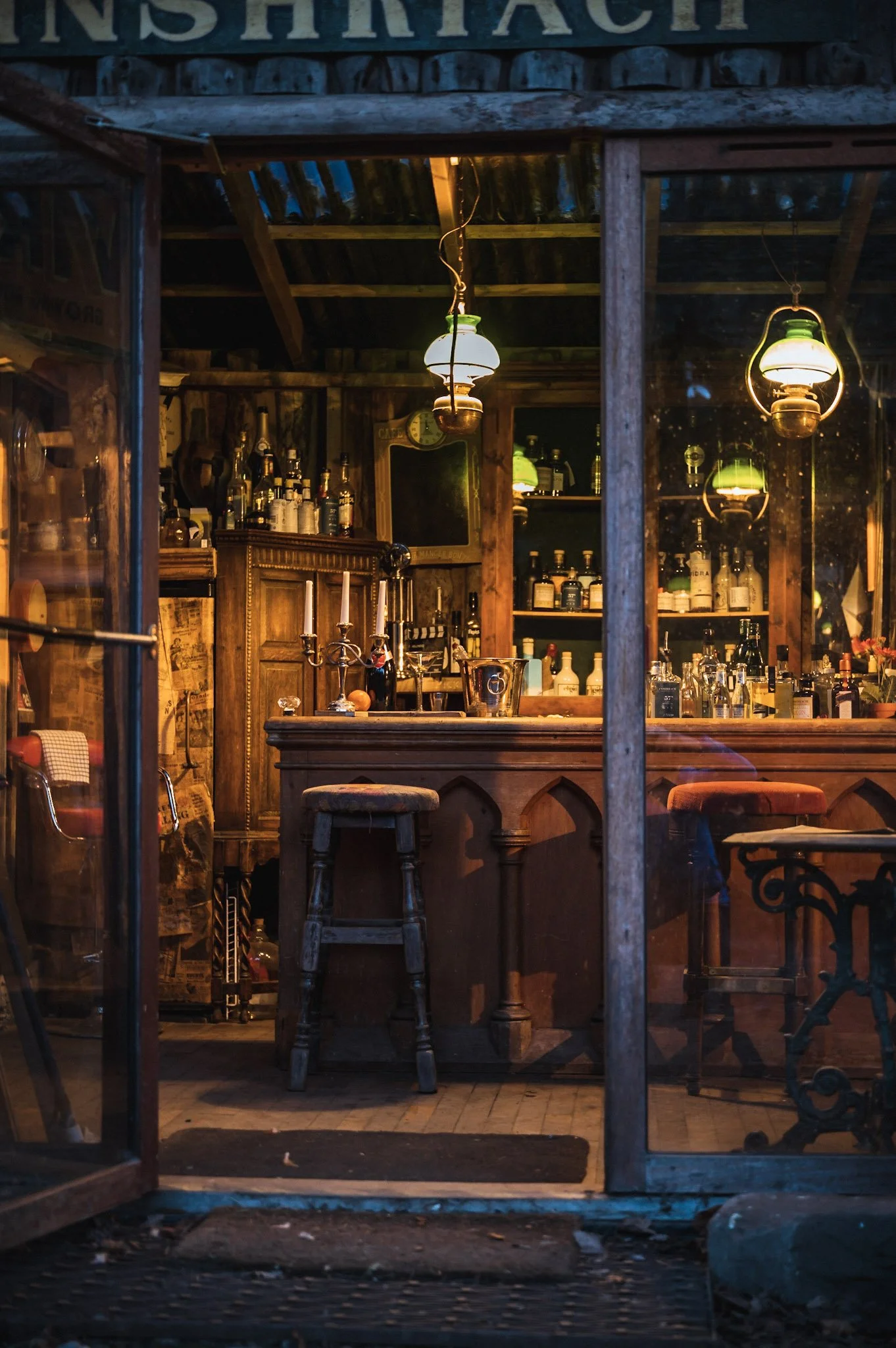 Inside a cozy, dimly lit rustic bar with wooden furniture, vintage lamps hanging from the ceiling, and bottles of liquor displayed on shelves.