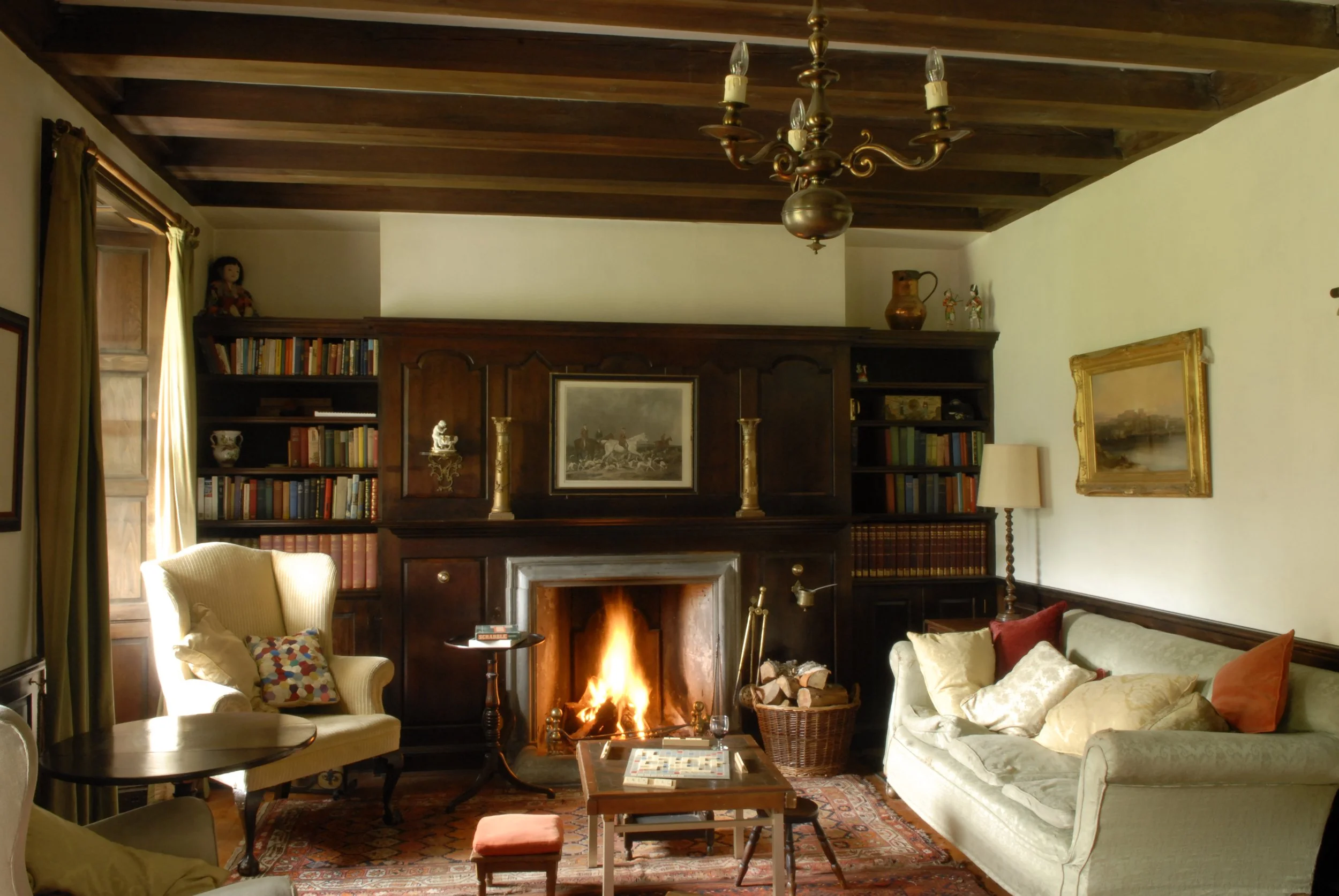 Living room with a fireplace, wooden ceiling beams, bookshelves, a chandelier, a lamp, a sofa with pillows, an armchair, a small table with a game board, and an area rug.
