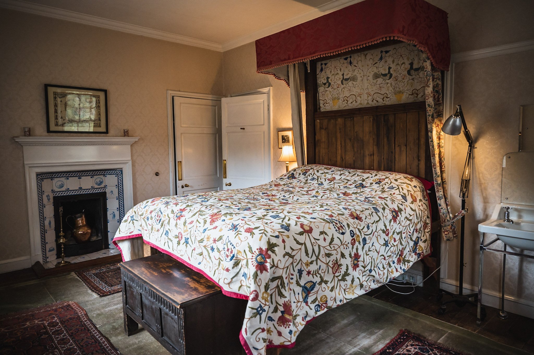 A bedroom with a four-poster bed featuring a wooden headboard, colorful floral quilt, and decorative curtains. There is a small fireplace with blue and white tiles under a picture frame, a bedside lamp, a floor lamp, a wooden chest at the foot of the bed, and a sink on the right side.