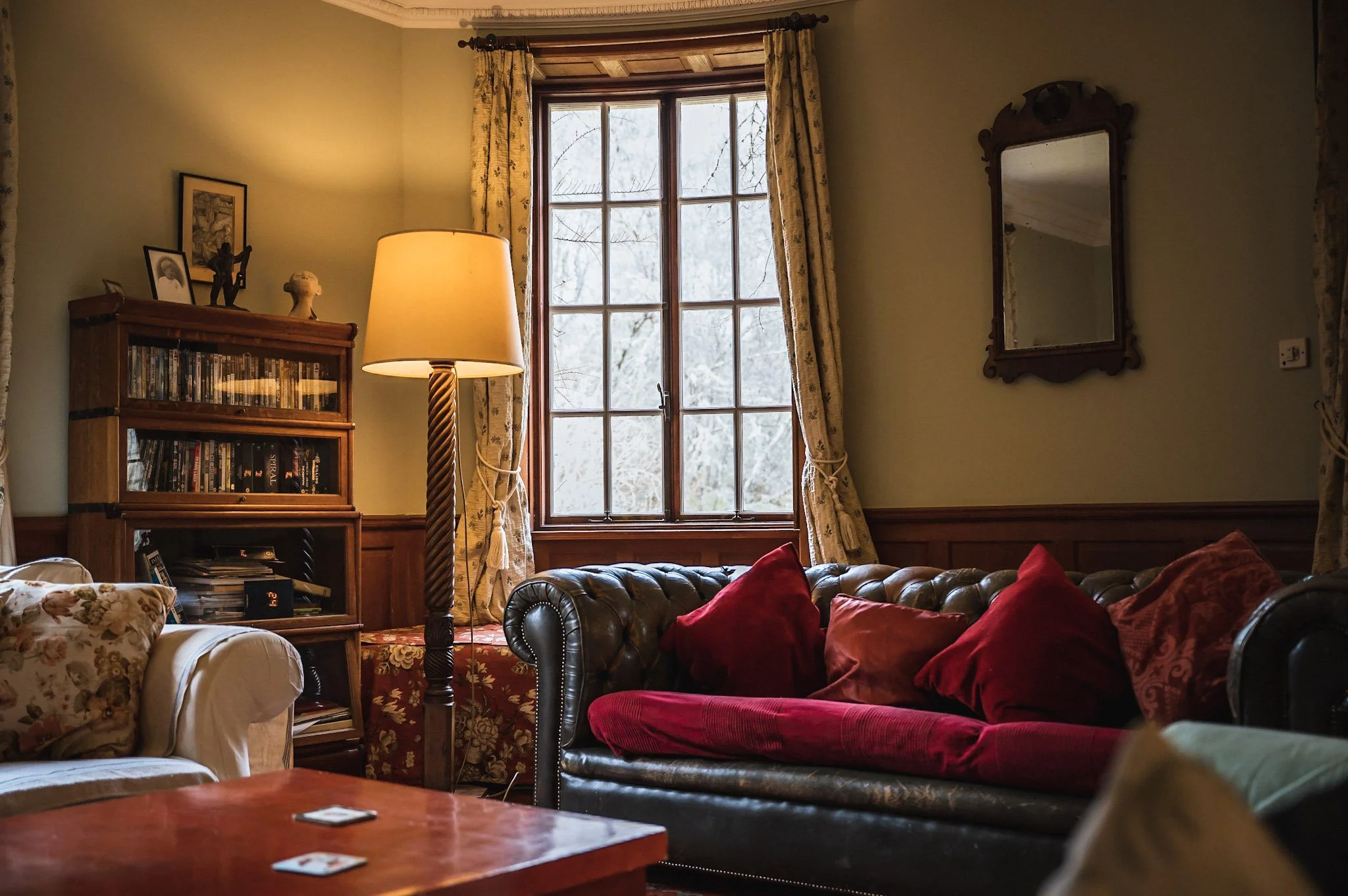 A cozy living room with a large window, floral curtains, a vintage brown leather sofa with red cushions, a wooden bookshelf filled with DVDs and books, a floor lamp, a floral armchair, and a wall mirror.