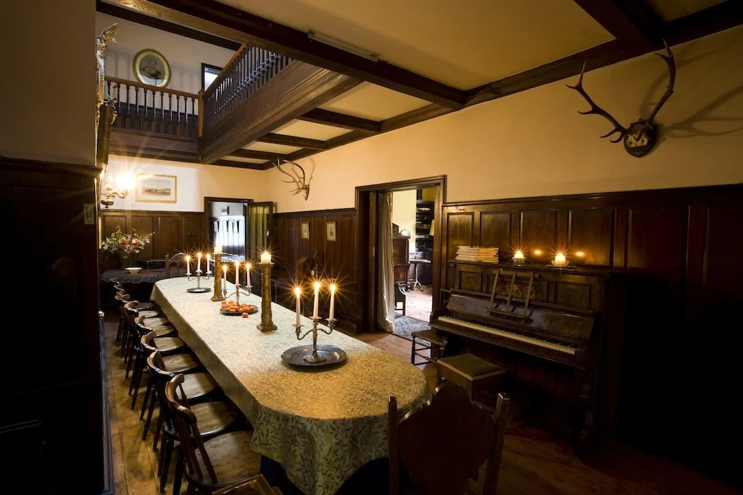 A dining room with a long table, lit by candles and surrounded by chairs. The room features wood-paneled walls, a piano, and mounted antlers.
