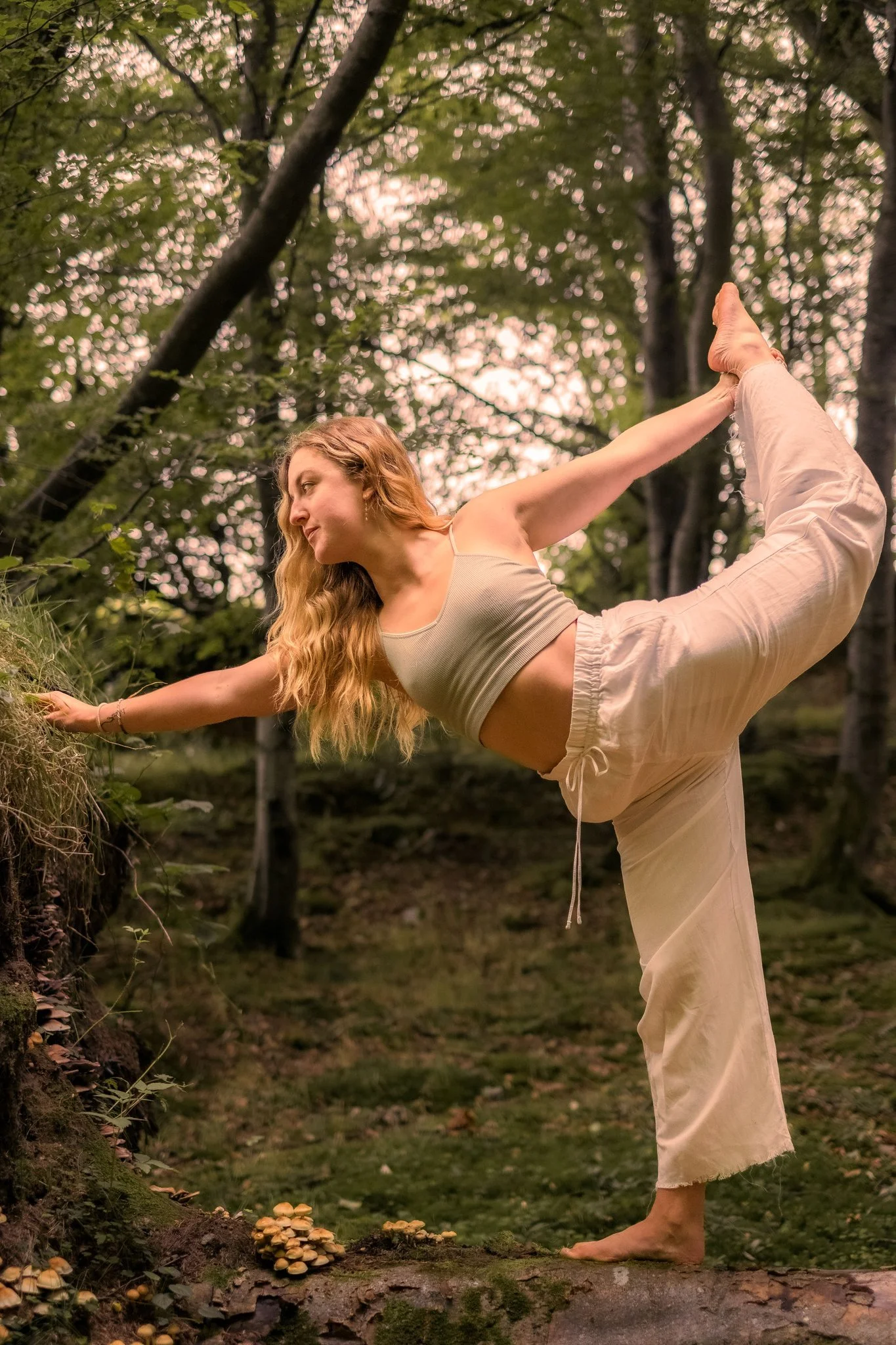 A woman practicing yoga outdoors on a mossy log in a forest, balancing in a dancer's pose with one leg lifted and her foot held by her hand.