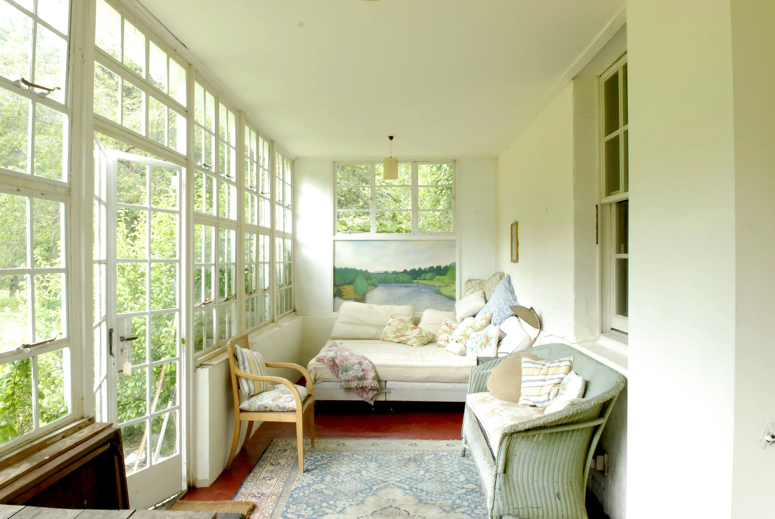 Sunlit enclosed porch with large windows, floral and wicker furniture, a painting of a river landscape, and a cozy cushioned seating area.