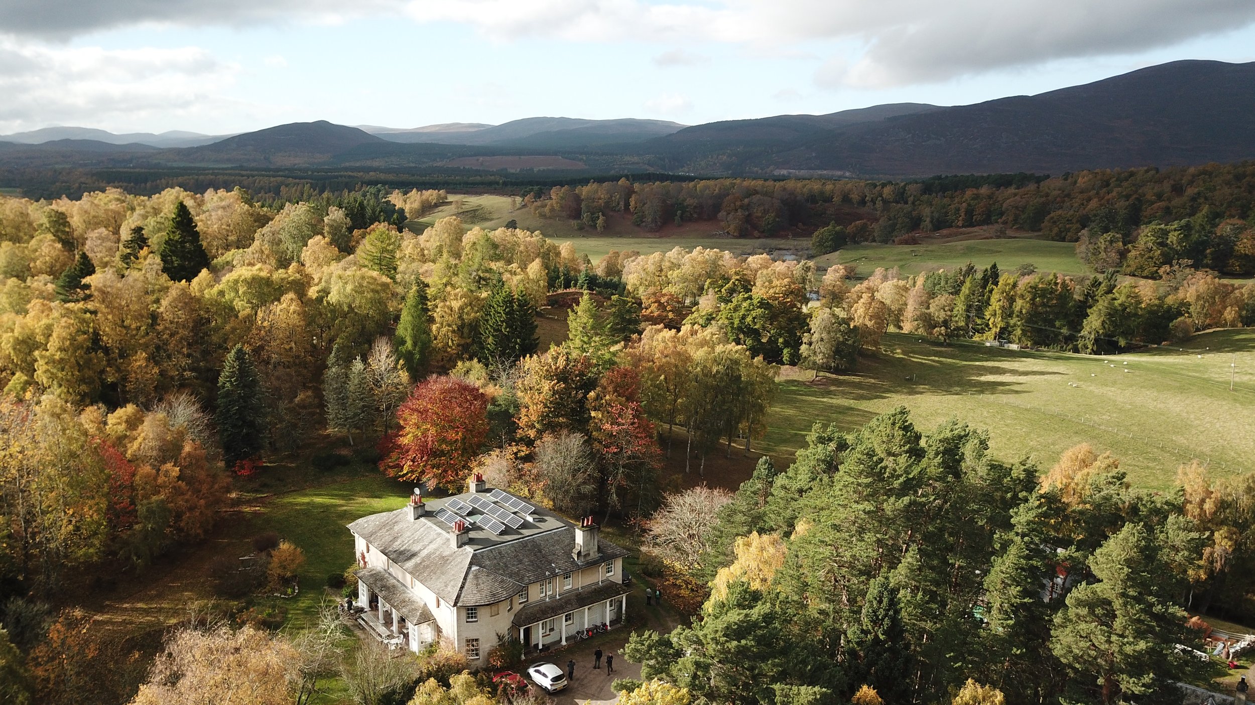 Aerial view of a large house with solar panels on the roof, surrounded by colorful autumn trees, in a mountainous landscape with hills and forests in the background.