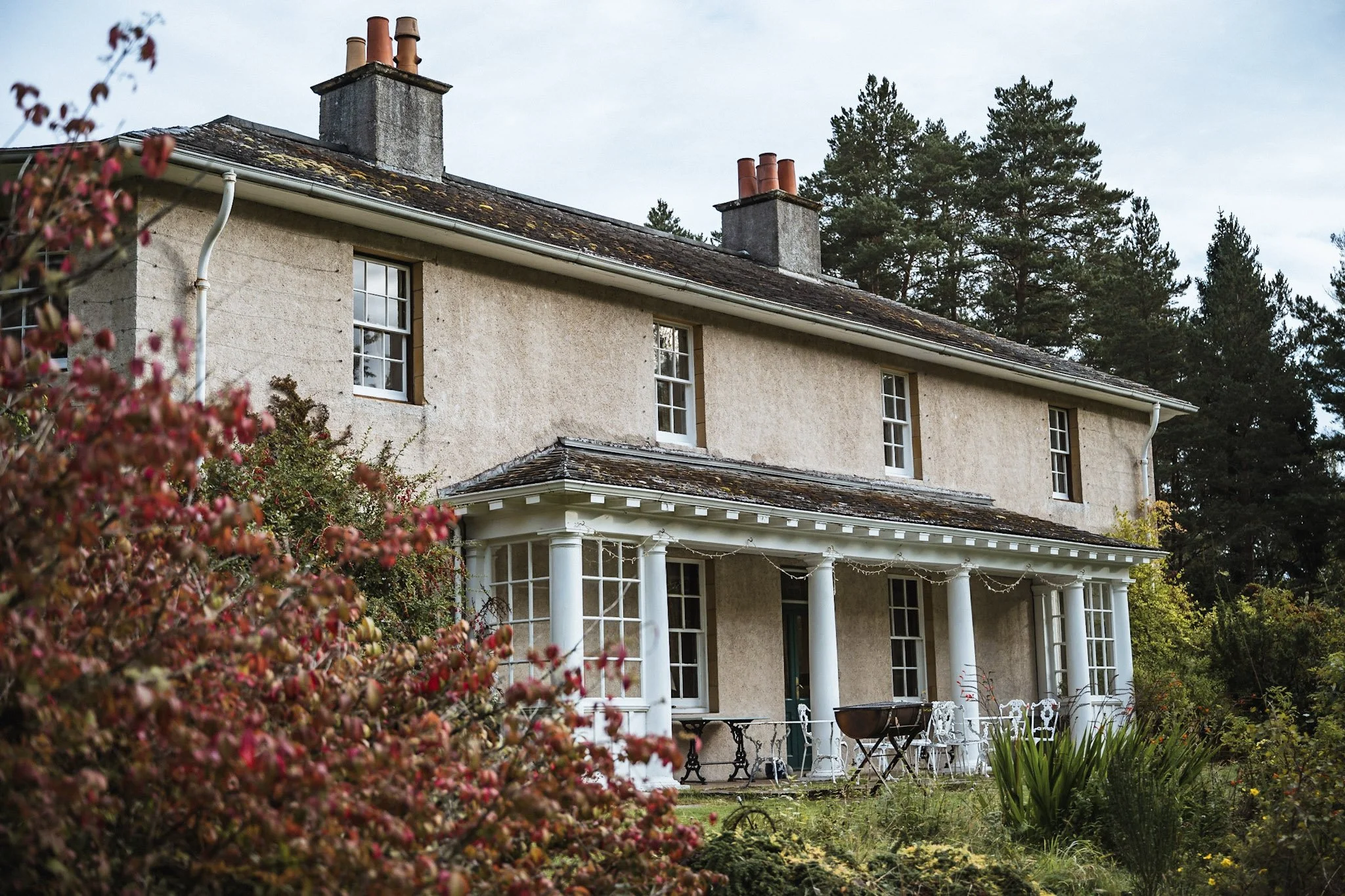 A two-story house with a beige exterior, multiple white-framed windows, and a covered porch with white columns. The roof has moss and lichen, and there are three chimneys. The house is surrounded by trees and garden plants, with outdoor furniture and a grill on the porch.