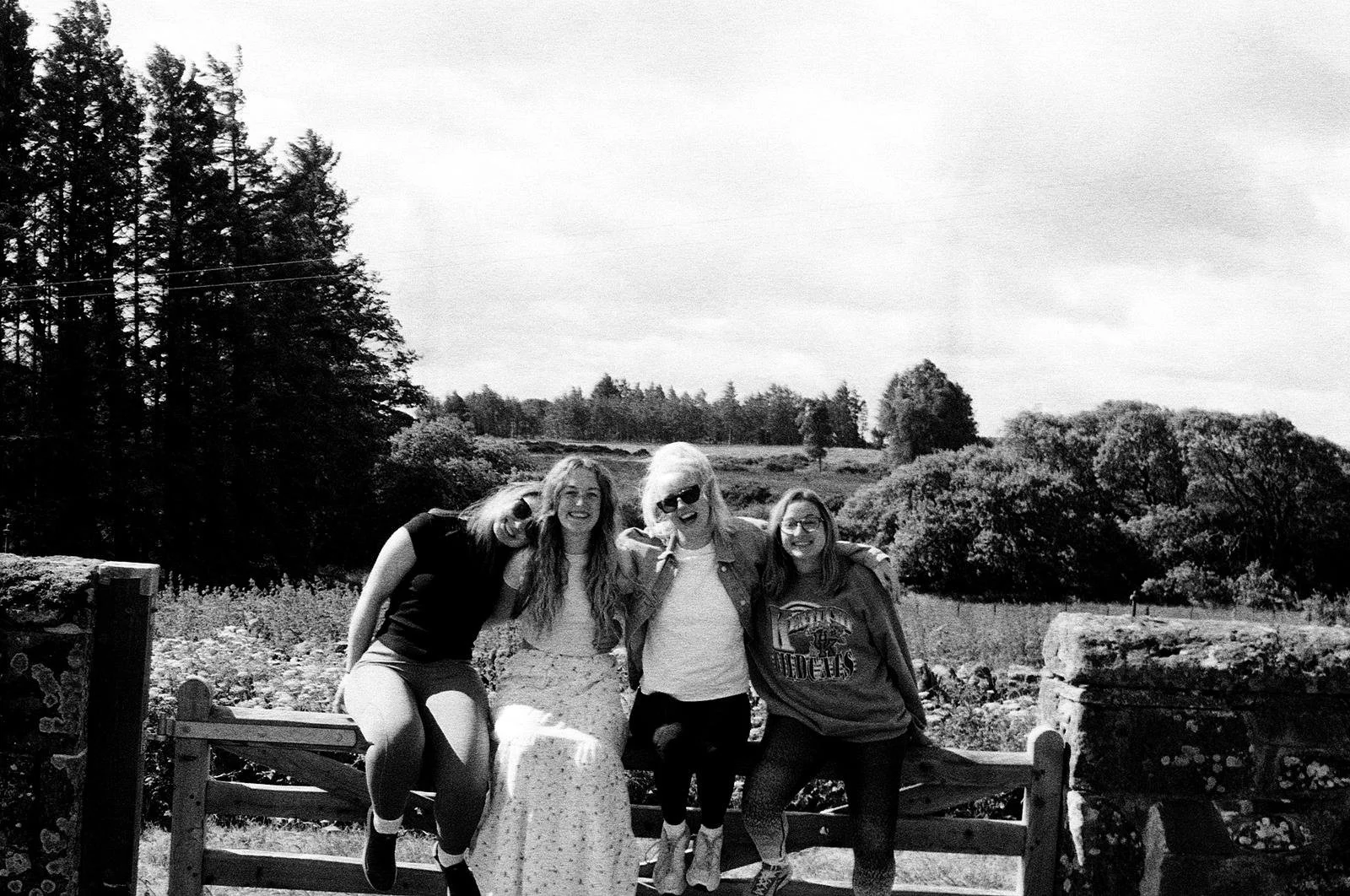 Four women sitting on a wooden fence outdoors, smiling at the camera with a scenic landscape of trees and open sky in the background.